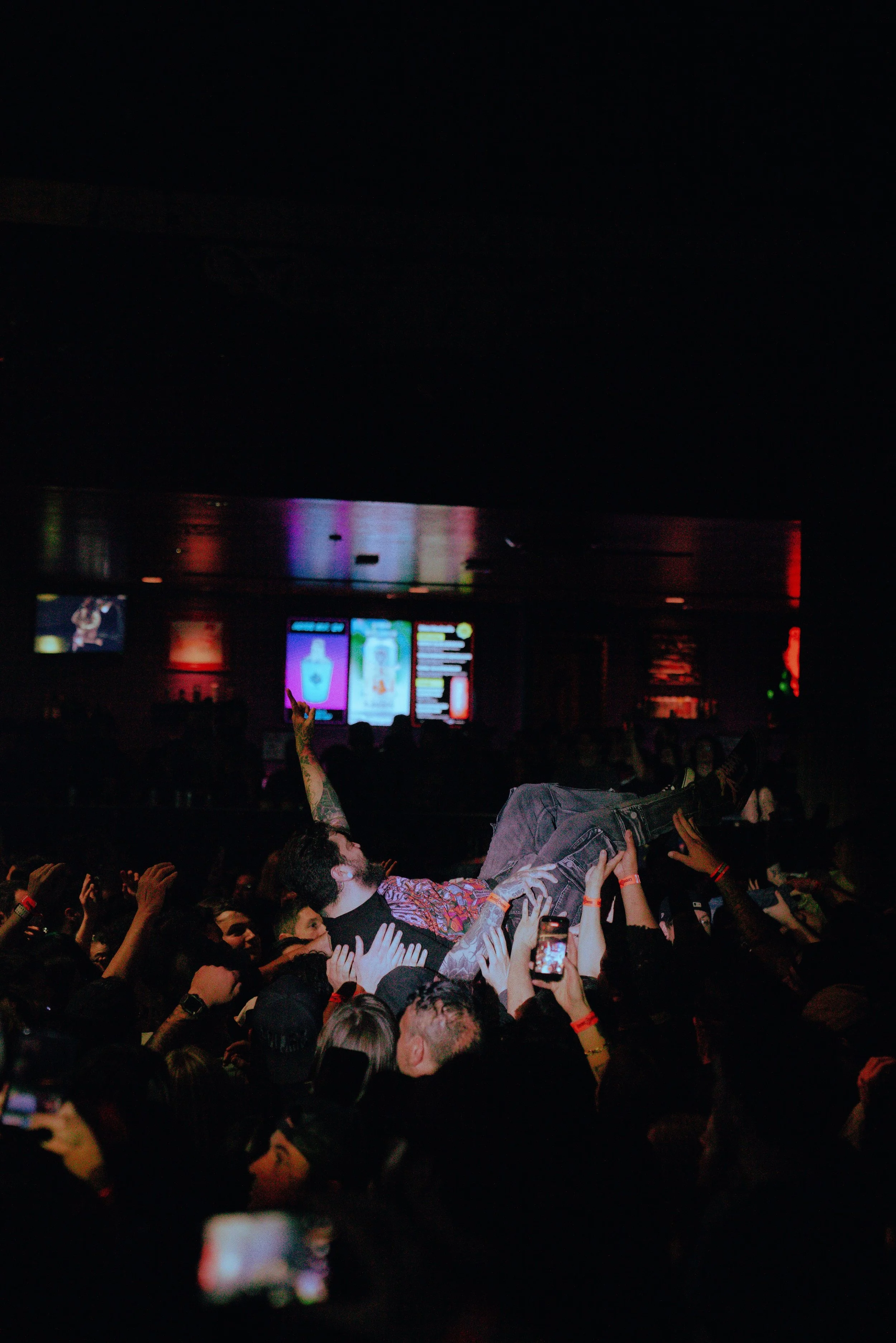 Crowd at concert or festival, with a person crowd surfing, held up by others in the audience, in a dark venue with colorful displays on screens.
