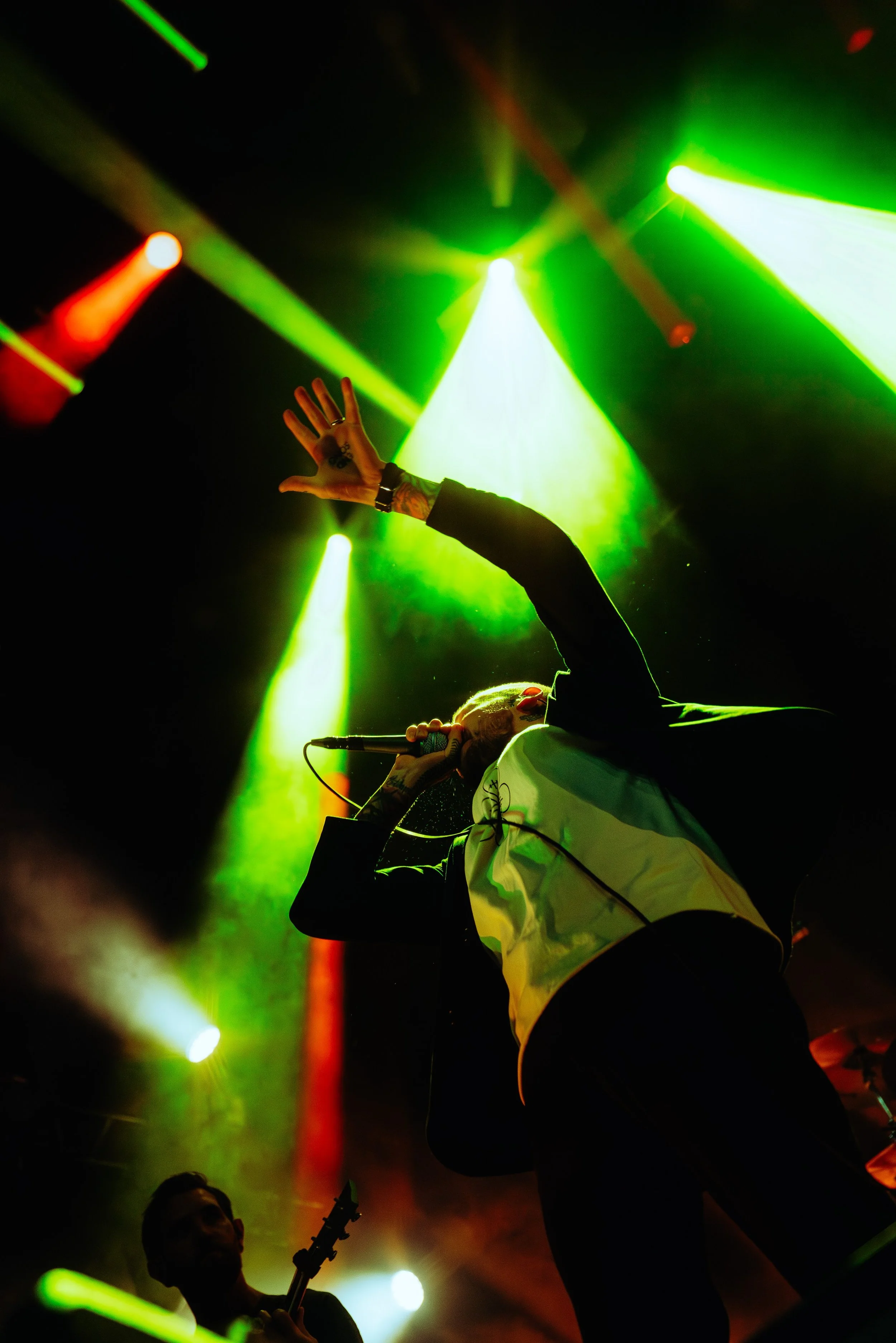 Performer singing into a microphone on stage with vibrant green and yellow stage lights, with a person playing guitar in the background.