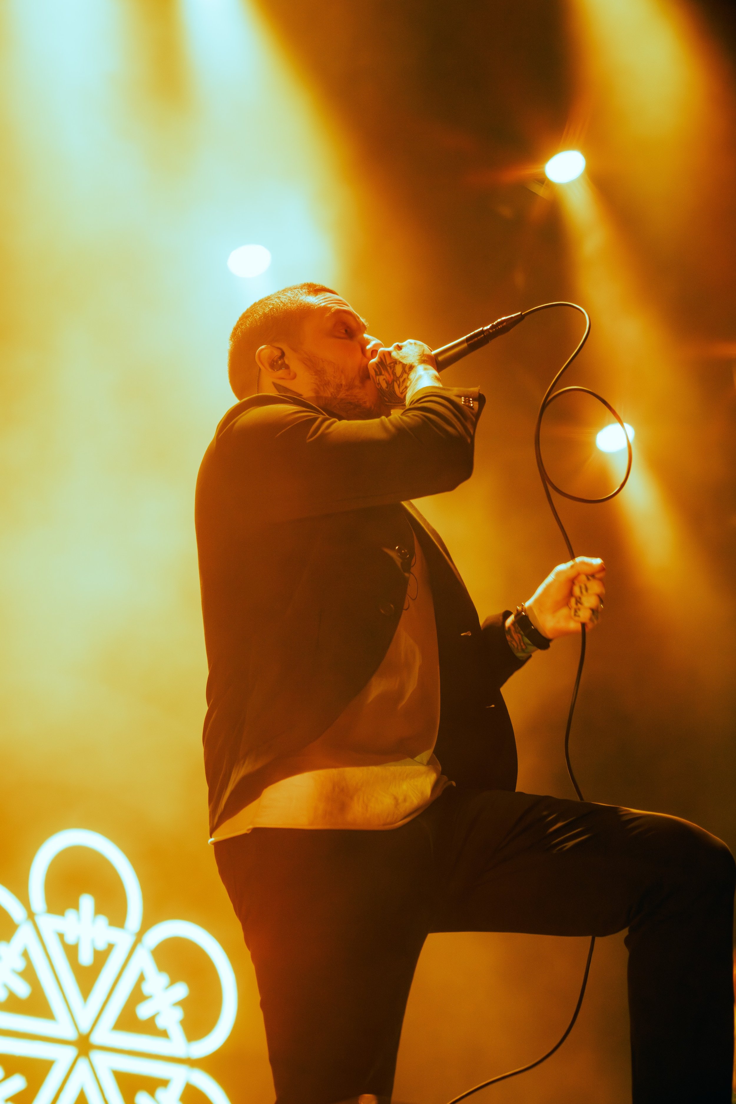 A singer performing on stage, holding a microphone with a yellow-orange background and illuminated decorative light structures.