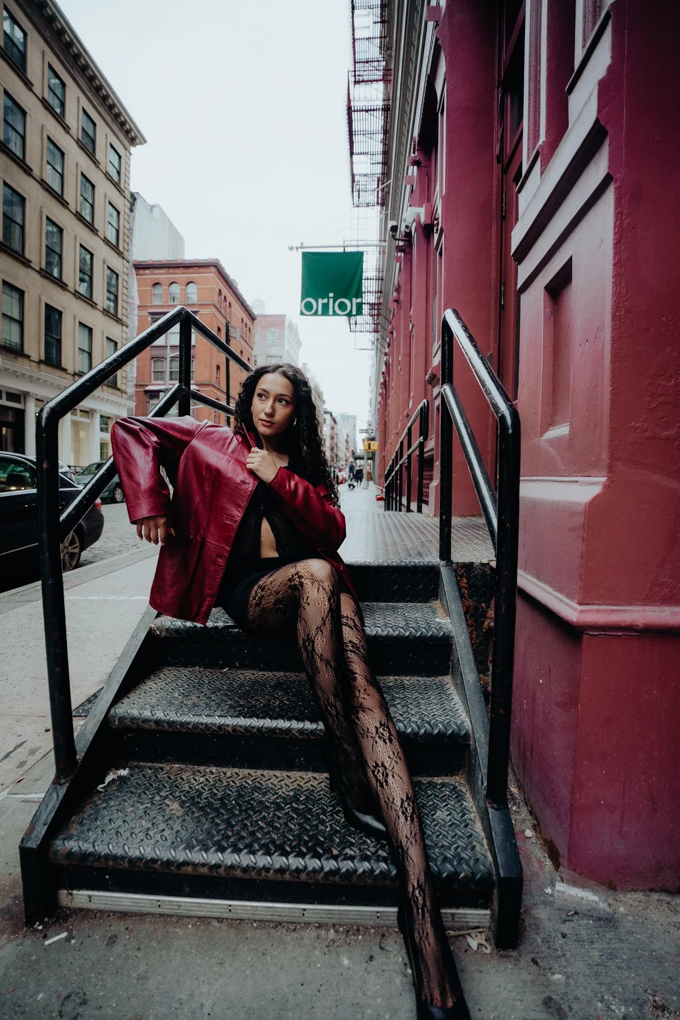 A woman with curly black hair sitting on outdoor stairs in an urban setting, wearing a red leather jacket, black dress, fishnet stockings, and black boots, with tall buildings in the background.