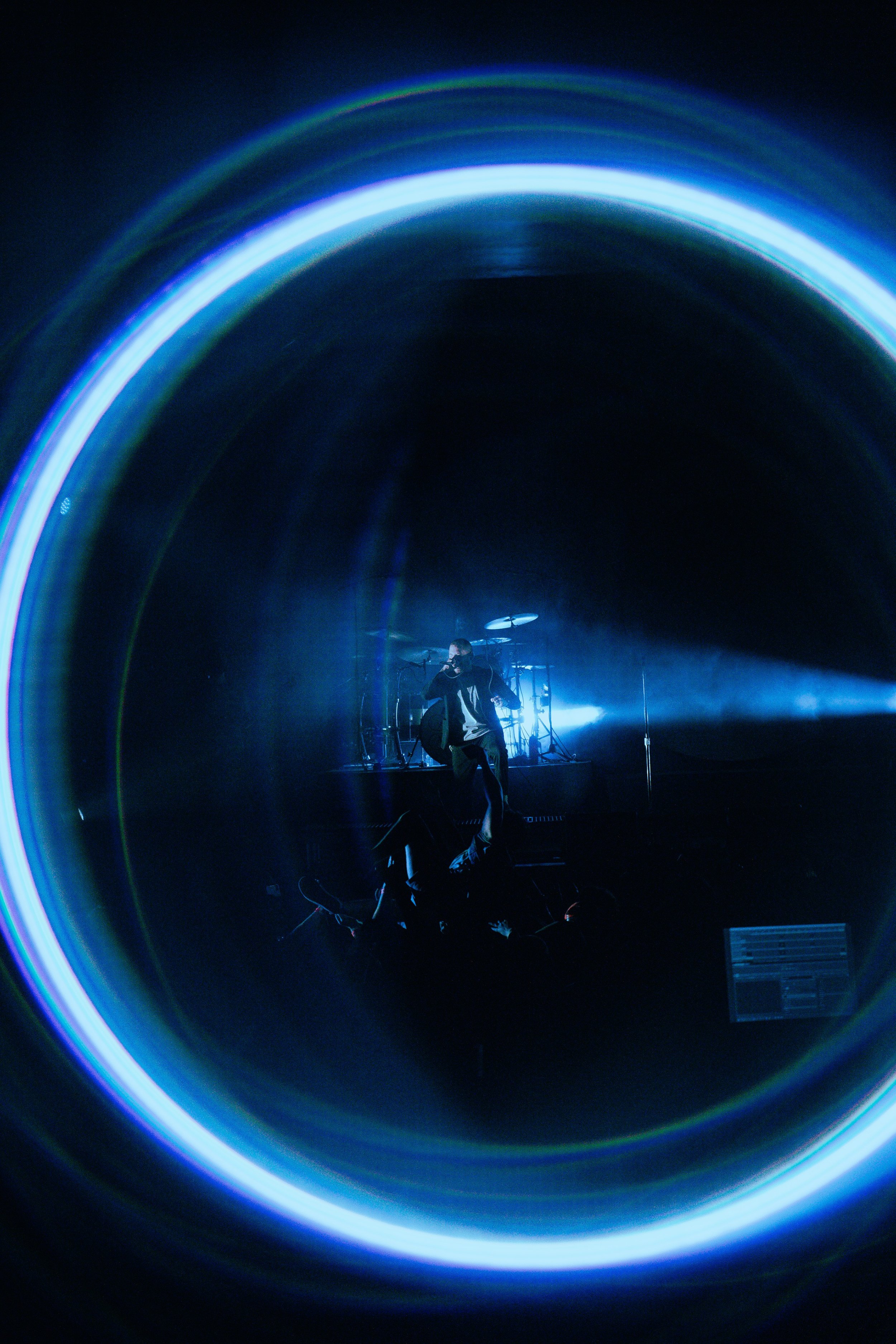 A performer on stage with a microphone, surrounded by vibrant light rings, performing in front of audience with a drum set in the background.