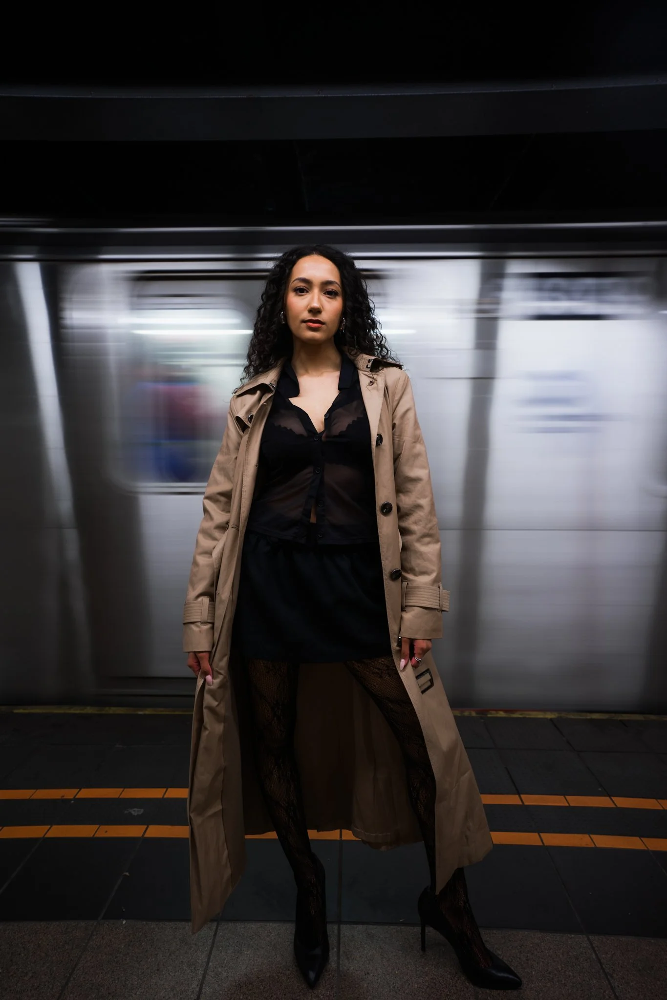 A woman standing with her hands at her sides in a subway station. The background shows a moving subway train with a metallic exterior and blurred windows. The woman has dark, curly hair, and wears a tan trench coat over black lace clothing, including