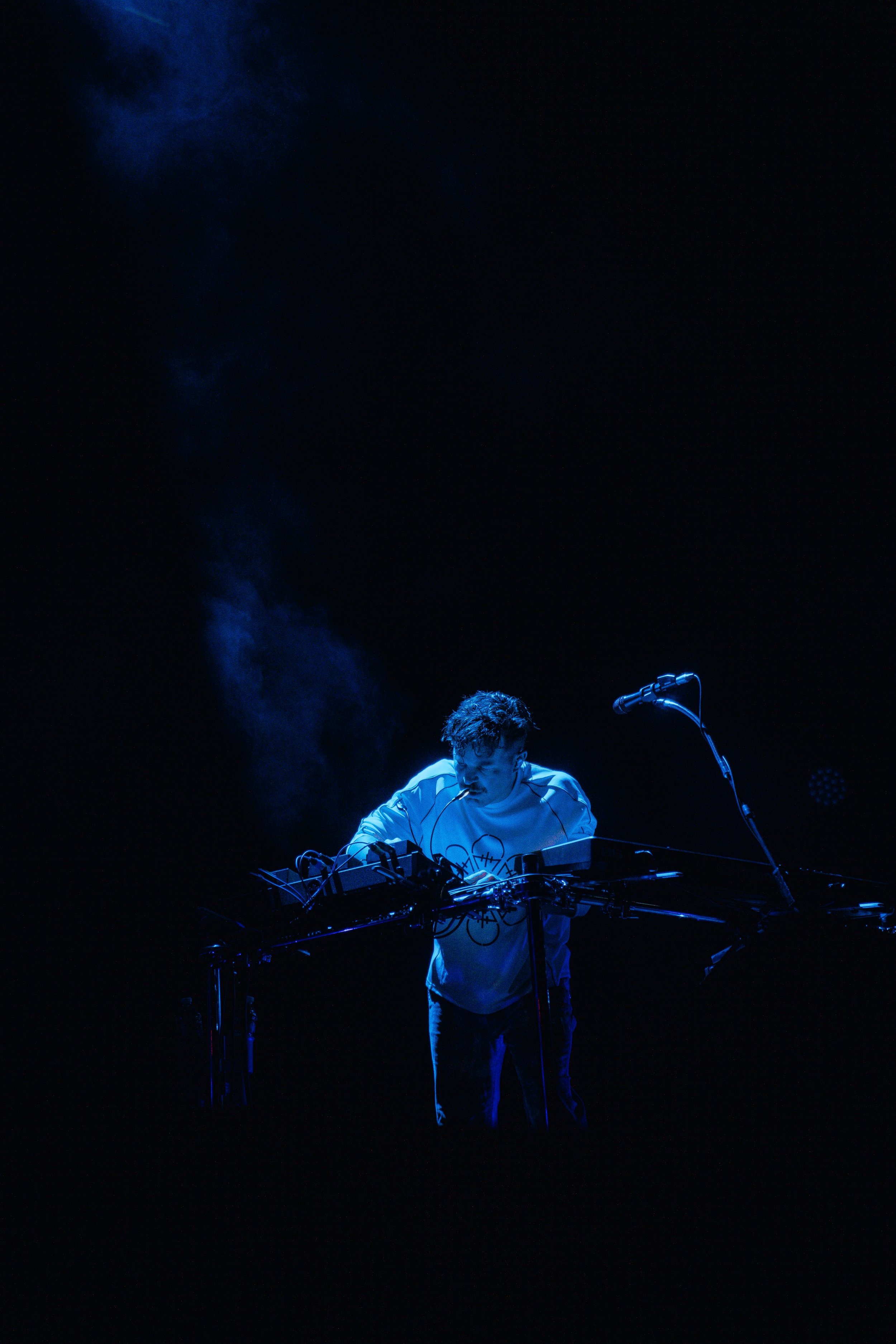 A person performing electronic music live on stage with various equipment, illuminated by blue stage lighting, against a dark background.