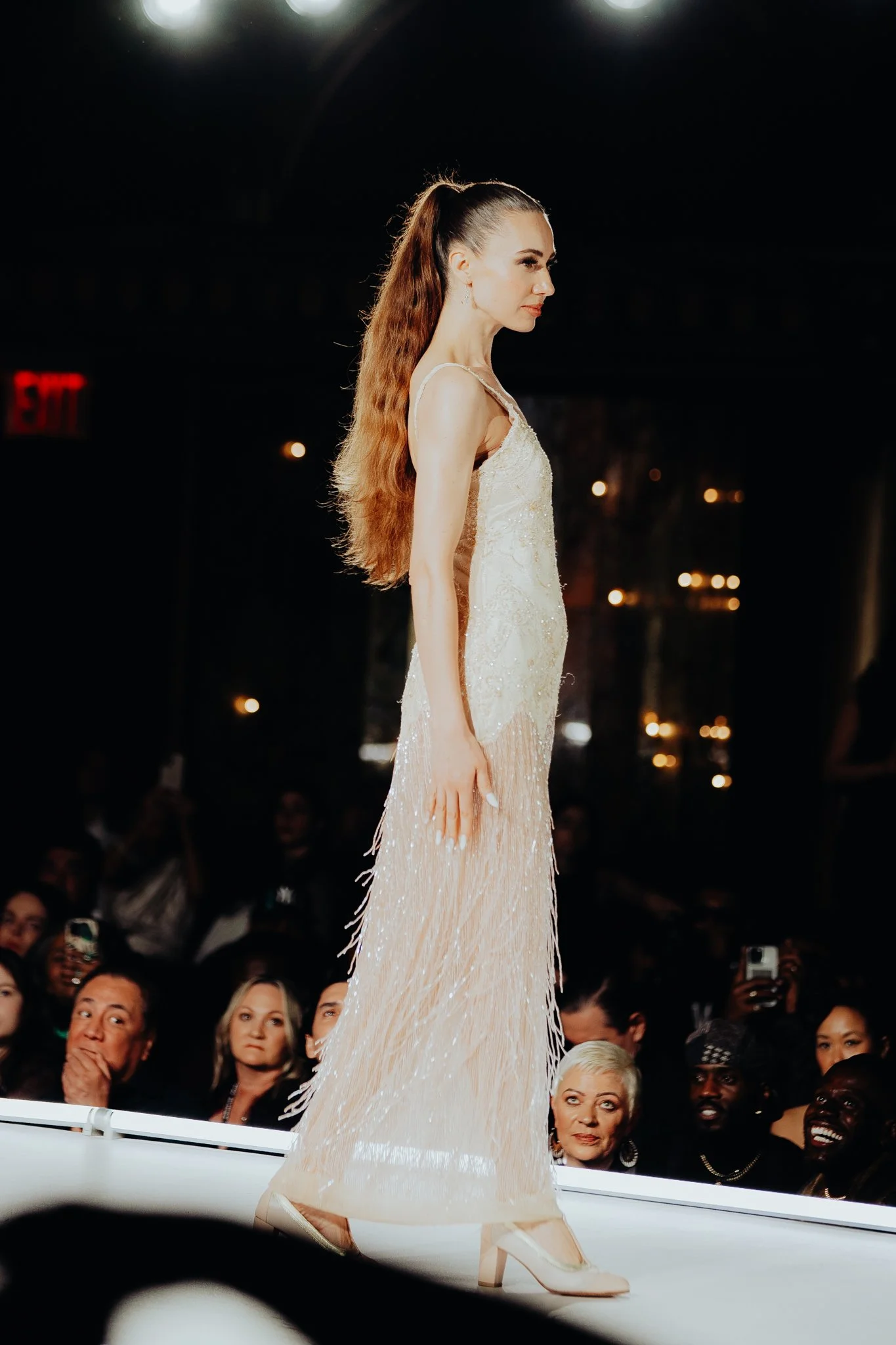 A model walking down a runway in a glamorous, light-colored gown with a high slit, paired with heels. The audience watches attentively, some taking photos, in a darkened venue.