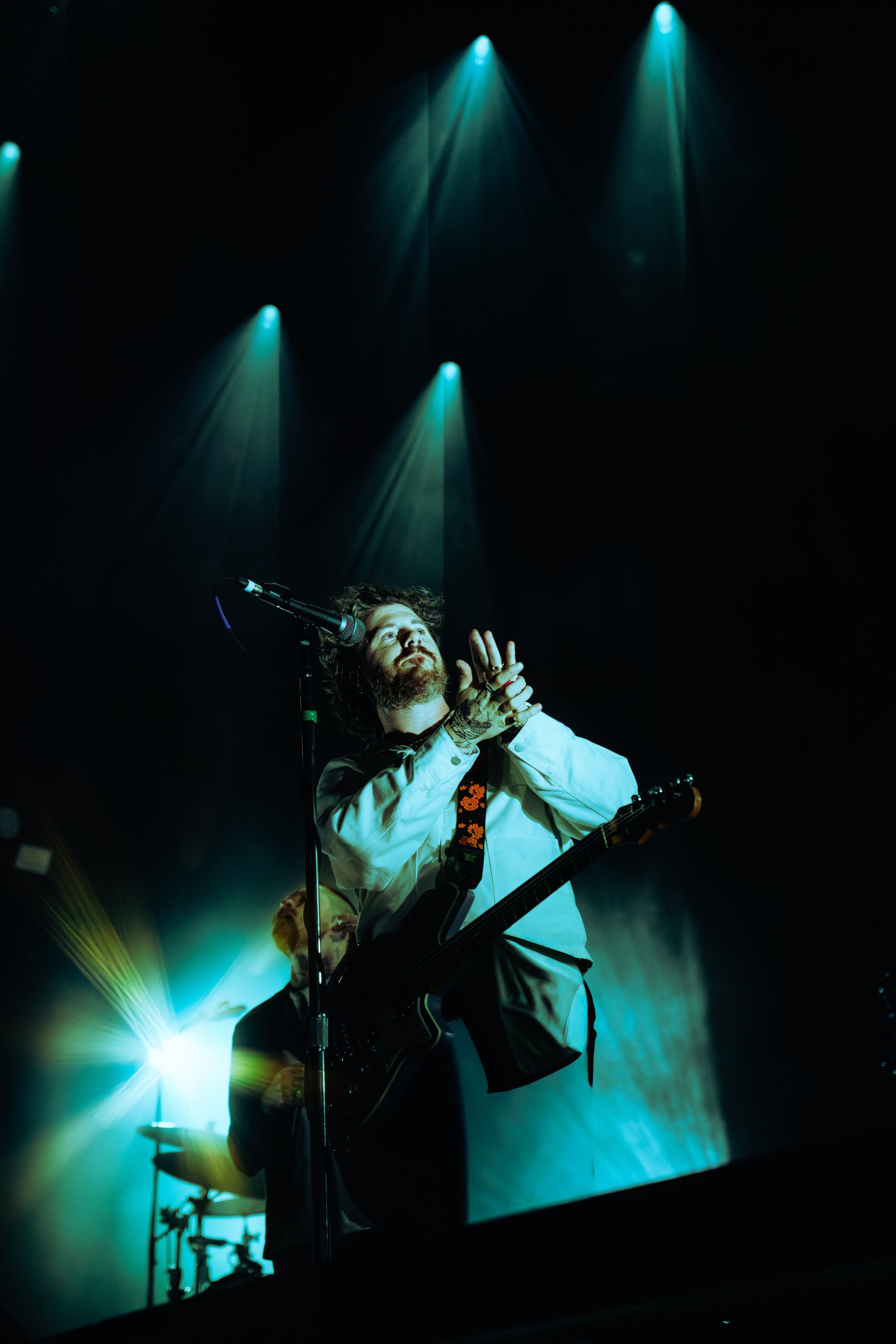 A male musician with a guitar performing on stage under blue and green spotlights, with a microphone in front of him.