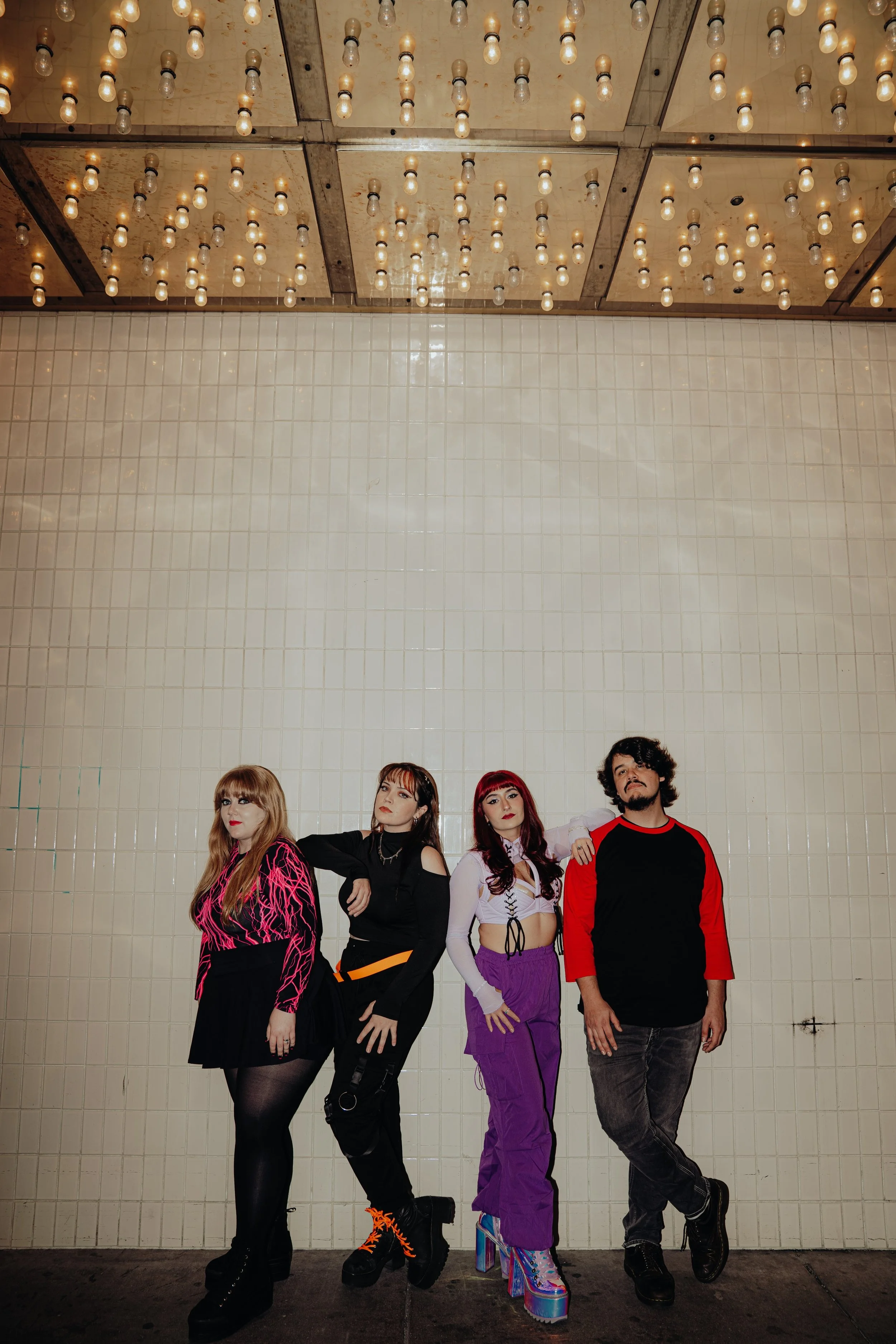 Five people standing against a tiled wall with ceiling lights above them. They are dressed in colorful, eclectic fashion.