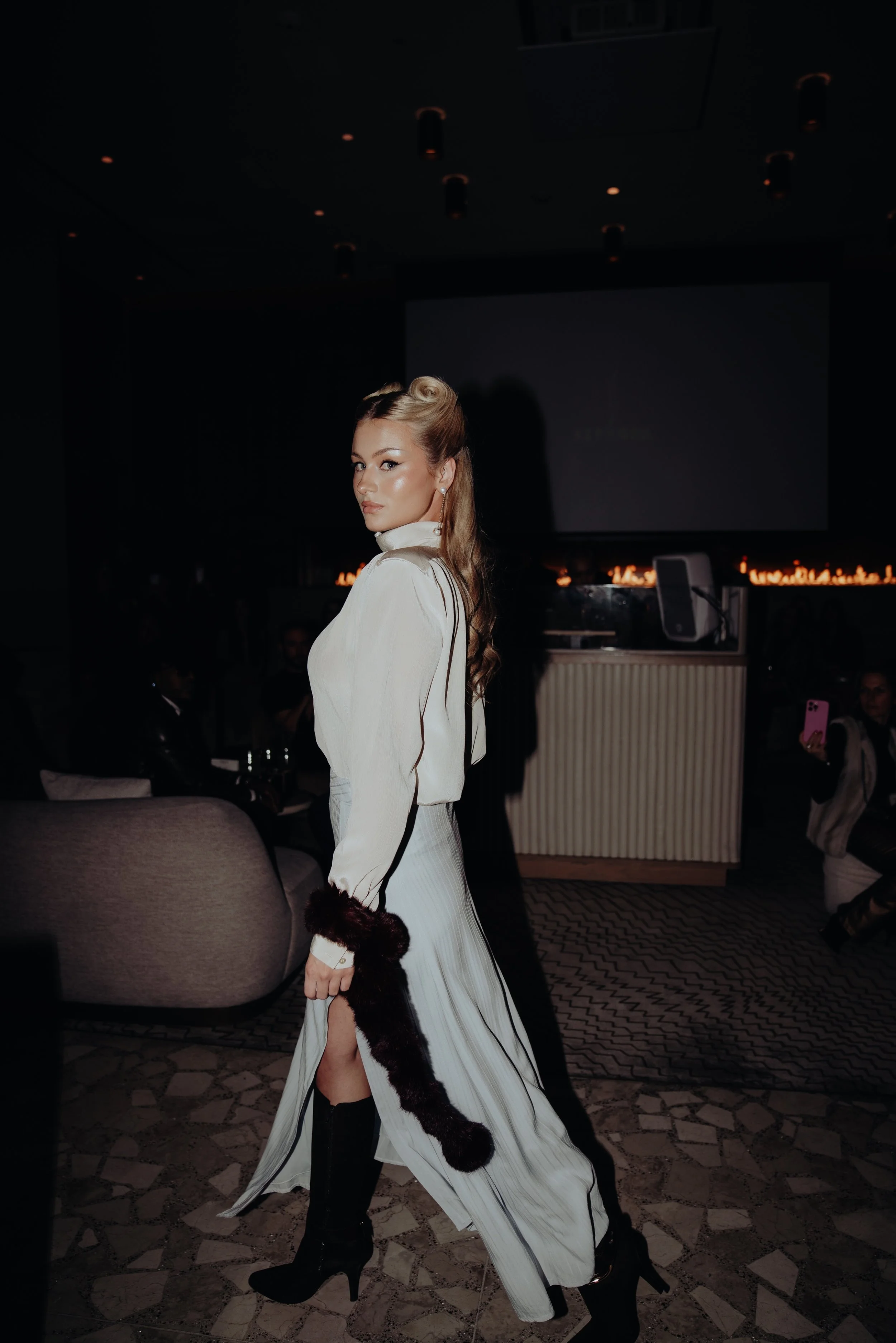 A woman with long blonde hair styled in vintage waves, wearing a white flowing dress with black accents and black high heels, stands in a dimly lit room with a dark background, capturing a portrait pose.