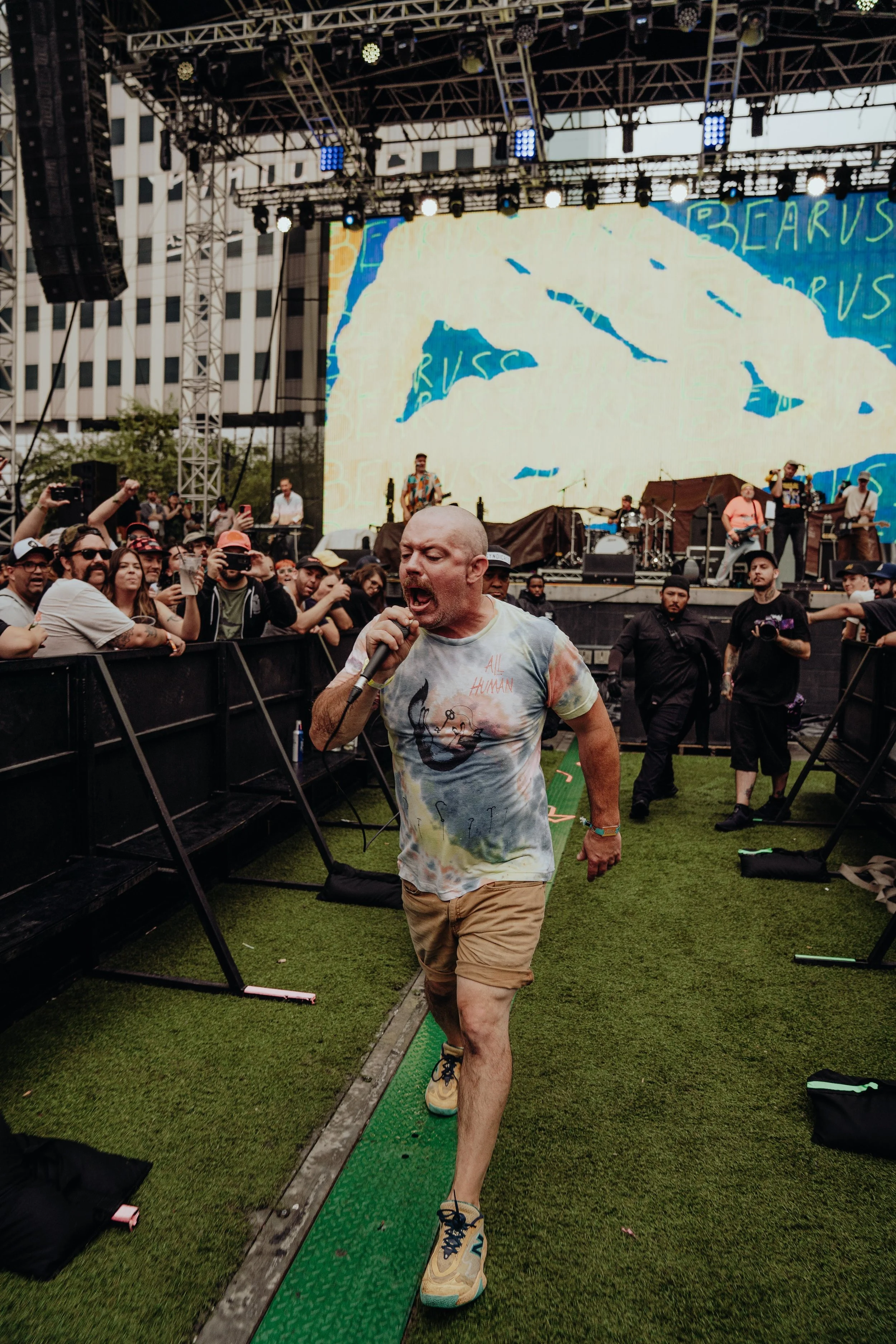 A man wearing a tie-dye shirt and beige shorts is walking on a green balancing platform at an outdoor concert event. He is holding a microphone close to his mouth, singing passionately. Behind him, there is a stage with musicians and a large screen d