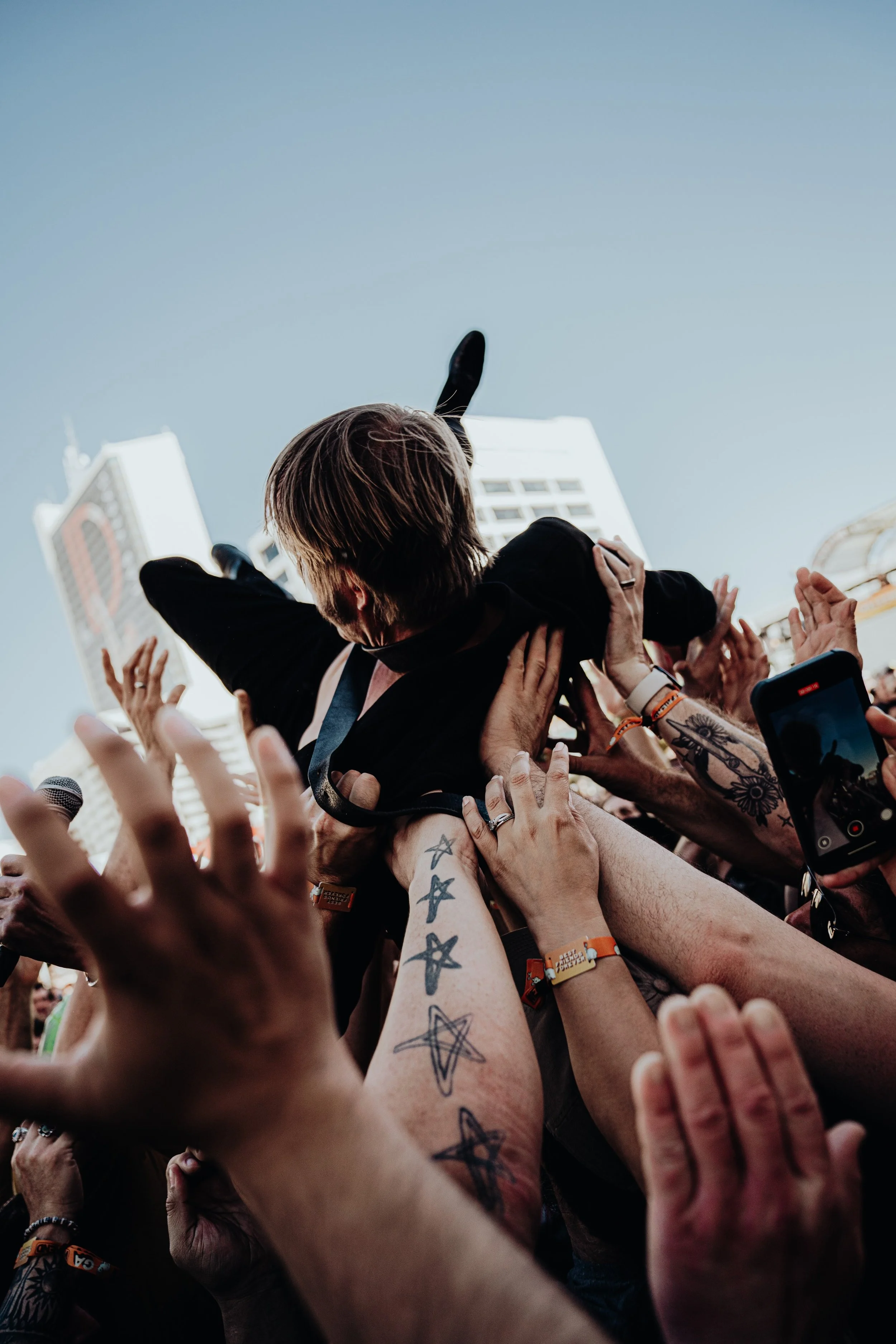 A crowd of people reaching up and supporting a person in a black jacket during an outdoor event with tall buildings in the background.