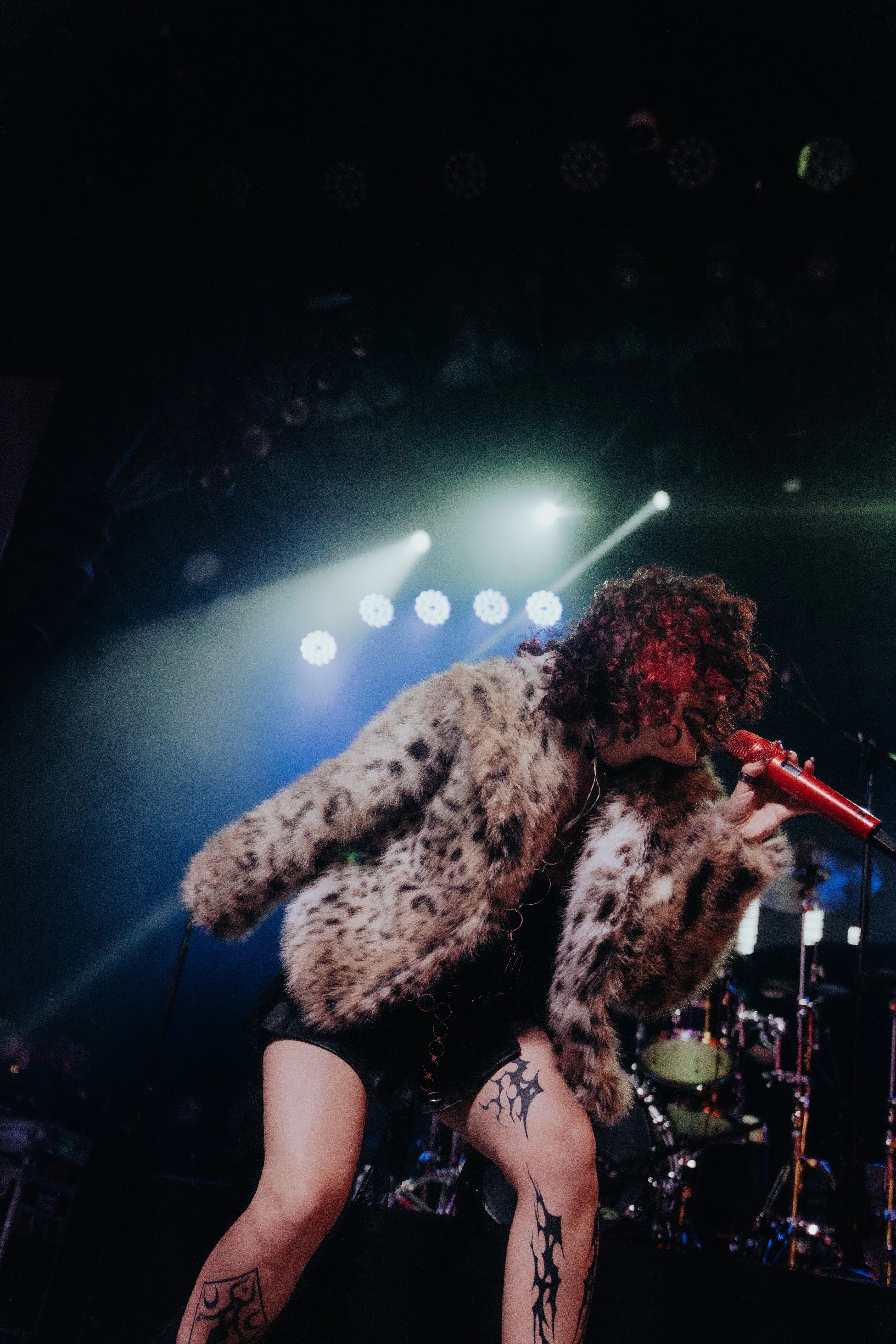 A female singer in a leopard print faux fur coat performing on stage, holding a microphone, with stage lights and music equipment in the background.