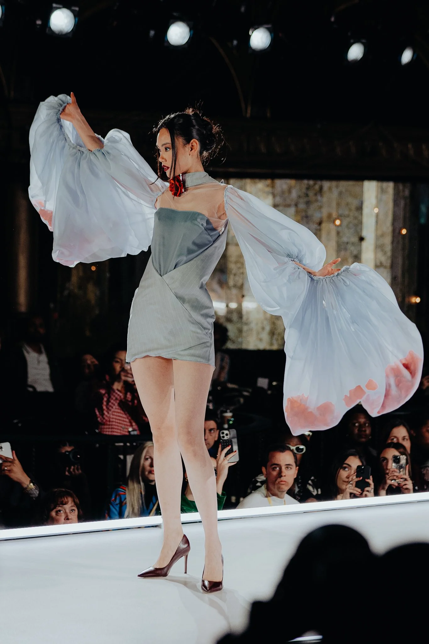 A woman in high heels performs on a runway, wearing a light gray dress with sheer, puffy white sleeves, surrounded by an audience of spectators.