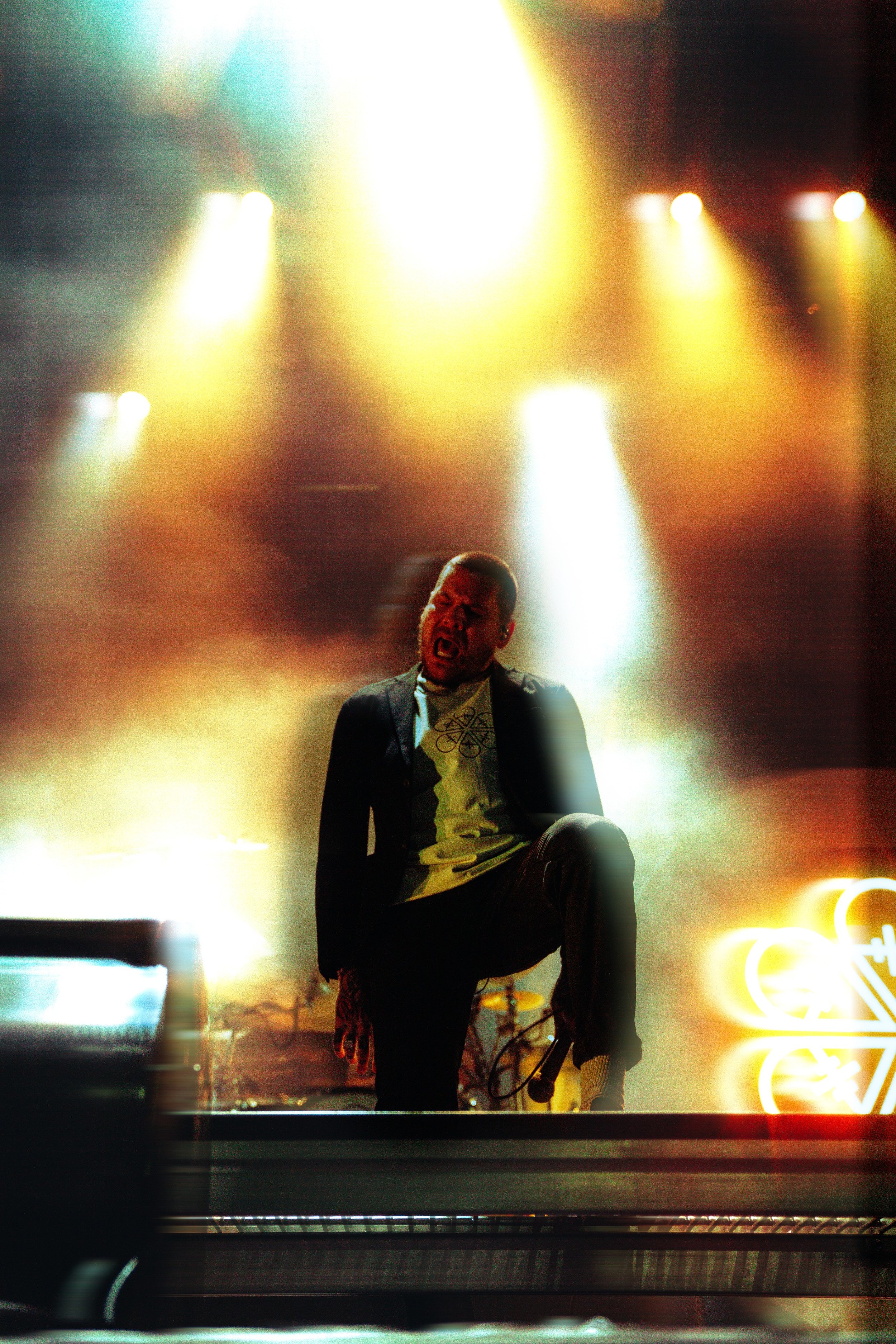 A man is singing passionately on stage under bright, multicolored stage lights. He is wearing a dark suit jacket over a T-shirt and is seated or crouched. The scene appears to be part of a concert or live performance with vibrant lighting effects.