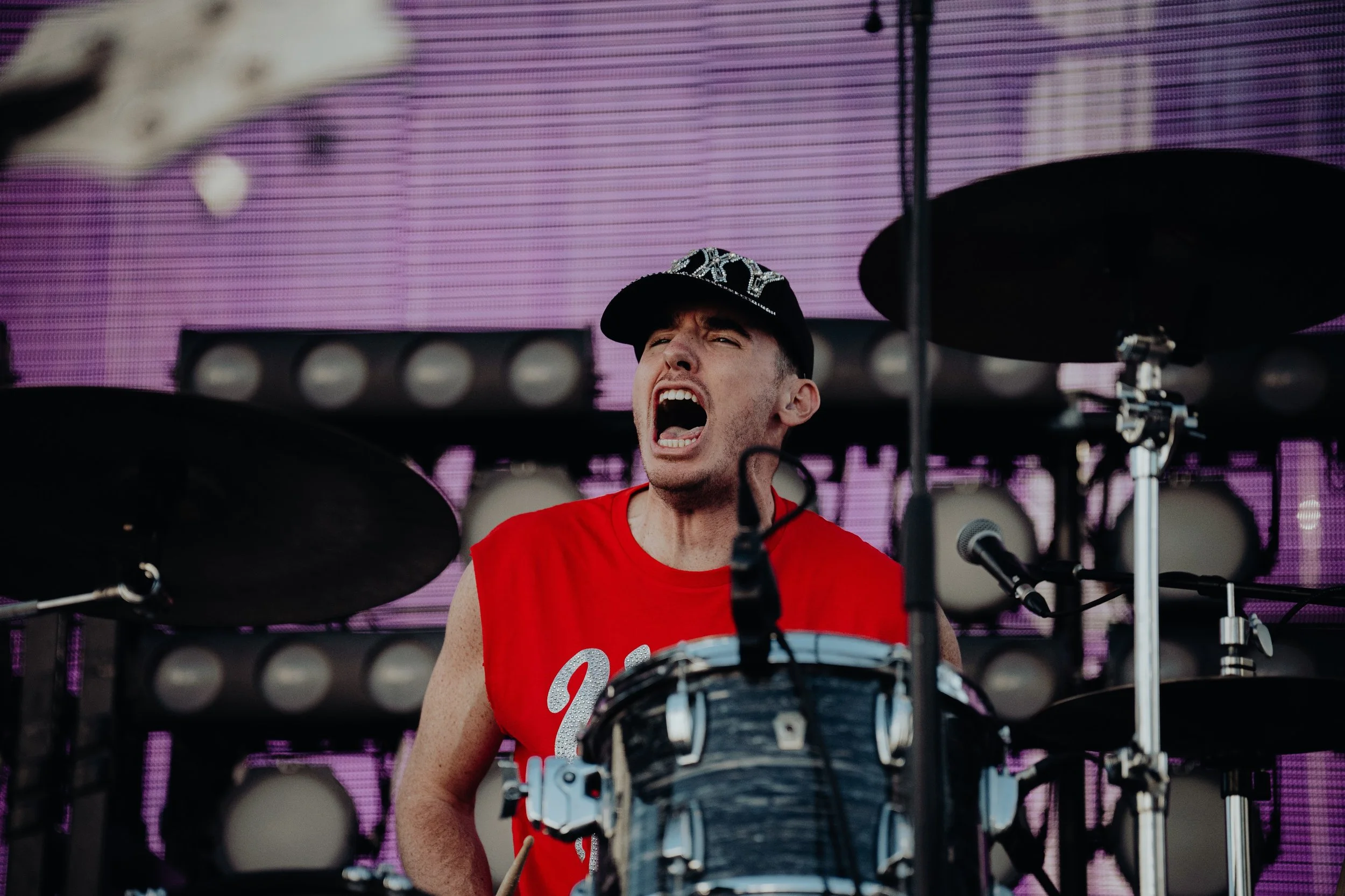 A young man with a snapback hat wearing a red sleeveless shirt, passionately singing or shouting on stage behind a drum set with a background of purple and pink lights.