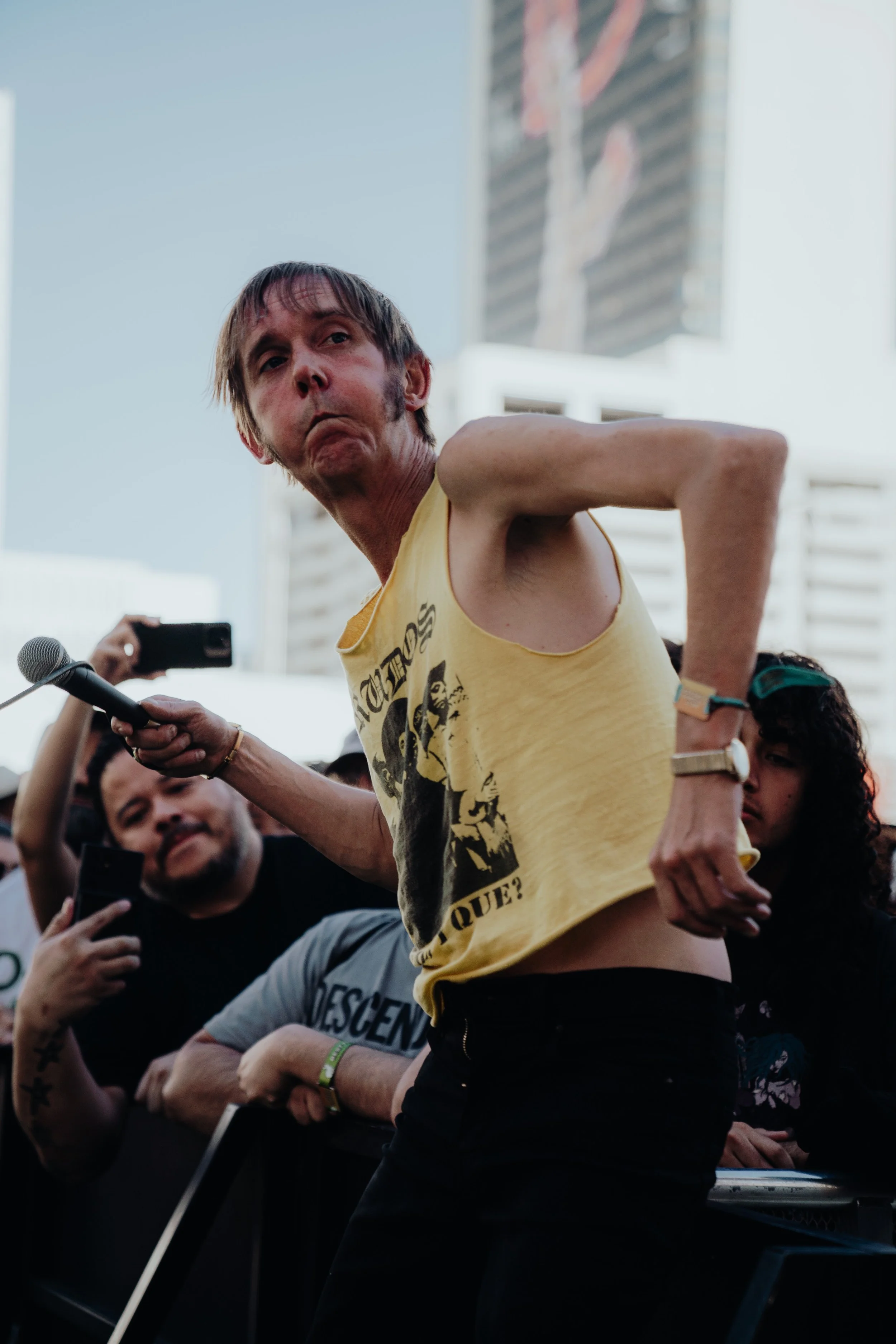A man with a yellow sleeveless shirt, with a serious expression, appears to be participating in a public event or protest in an urban setting, with buildings and several people around, some holding smartphones.