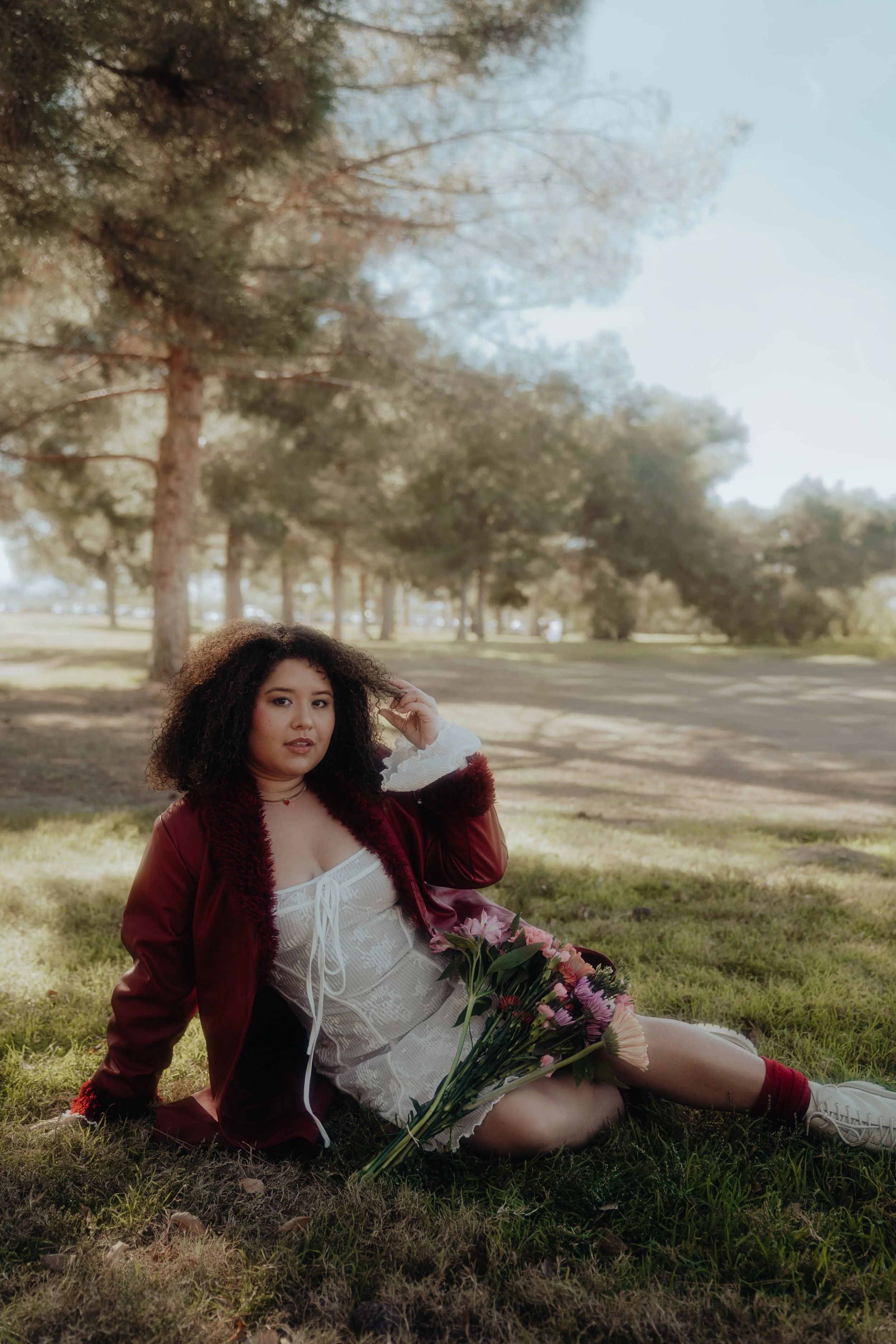A woman with curly hair sitting on grass in a park, holding a bouquet of flowers, dressed in a white dress and a red jacket, with a scenic background of trees and clear sky.