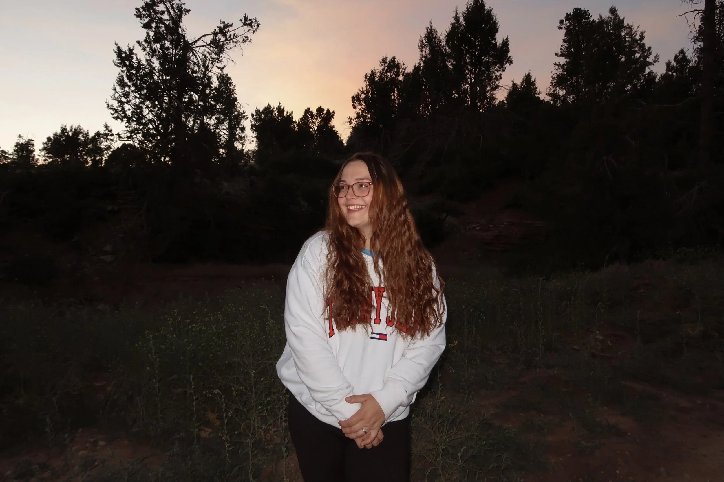 A woman with long, curly hair and glasses standing outdoors at dusk, smiling, with trees and a sunset sky in the background.