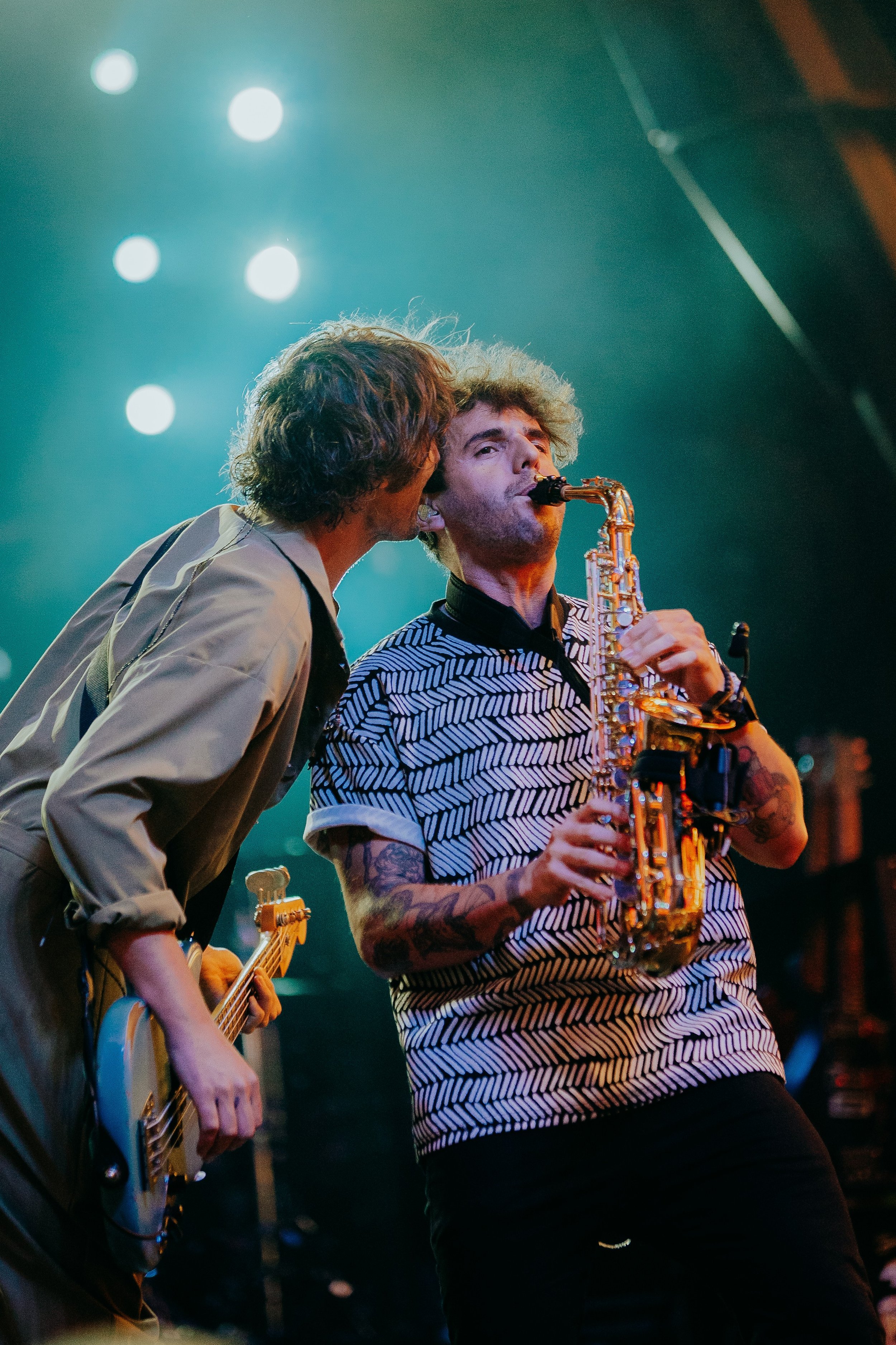 Two musicians performing on stage, one playing bass guitar and the other playing saxophone, with stage lights illuminating the scene.