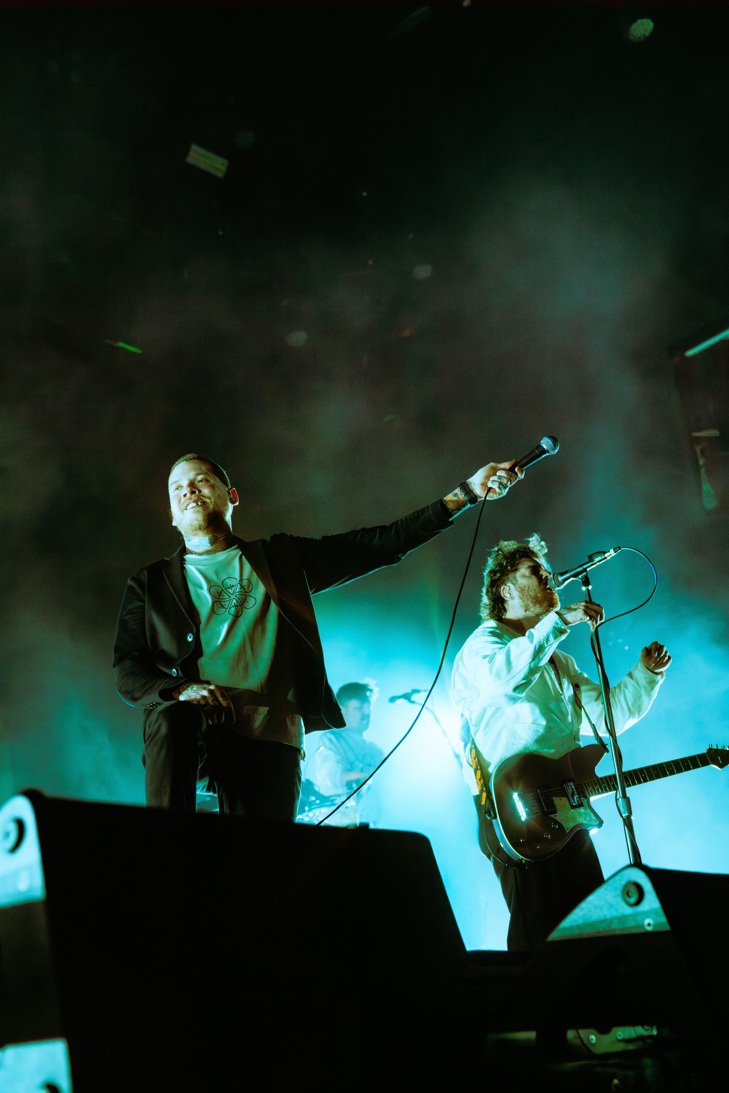 Musicians performing on stage with colorful lighting, one holding a microphone and another with a guitar.
