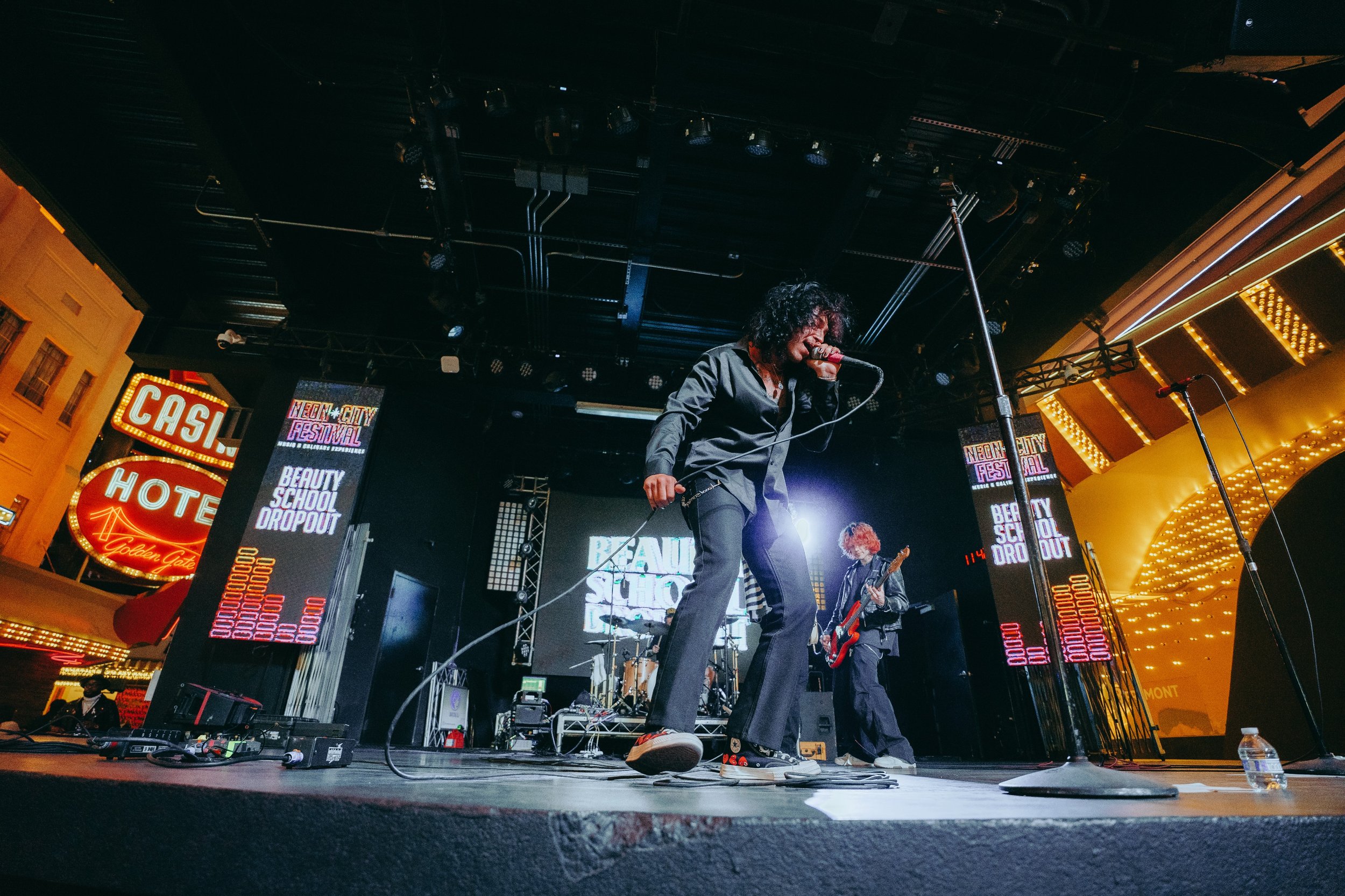 Musicians performing on stage during a Neon City Festival at night. Bright lights, signs, and a decorated building facade are visible, with one person singing into a microphone and others playing instruments.