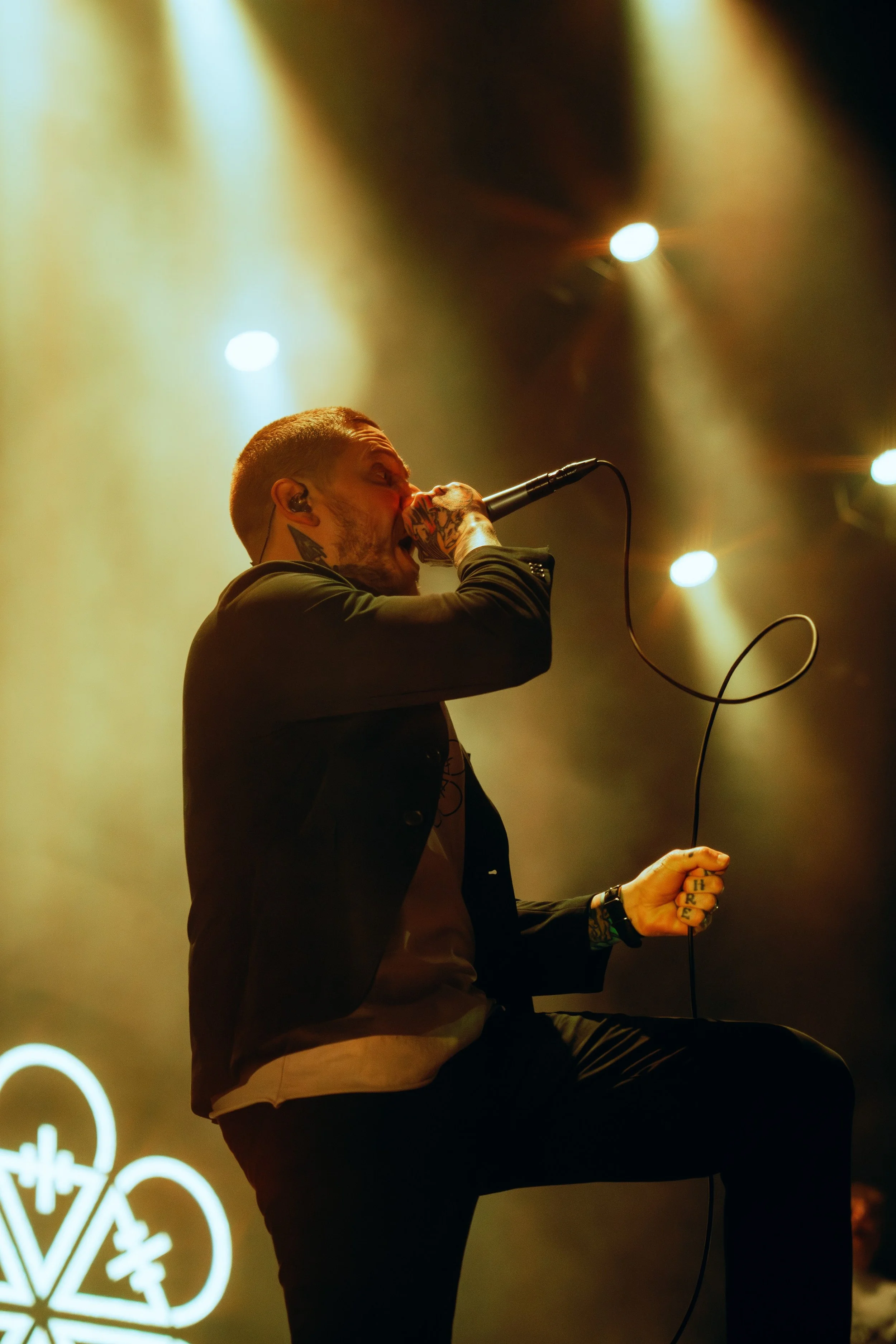 A male singer with tattoos on his neck and hand, performing on stage with a microphone in his hand, illuminated by stage lights and a blurred background.