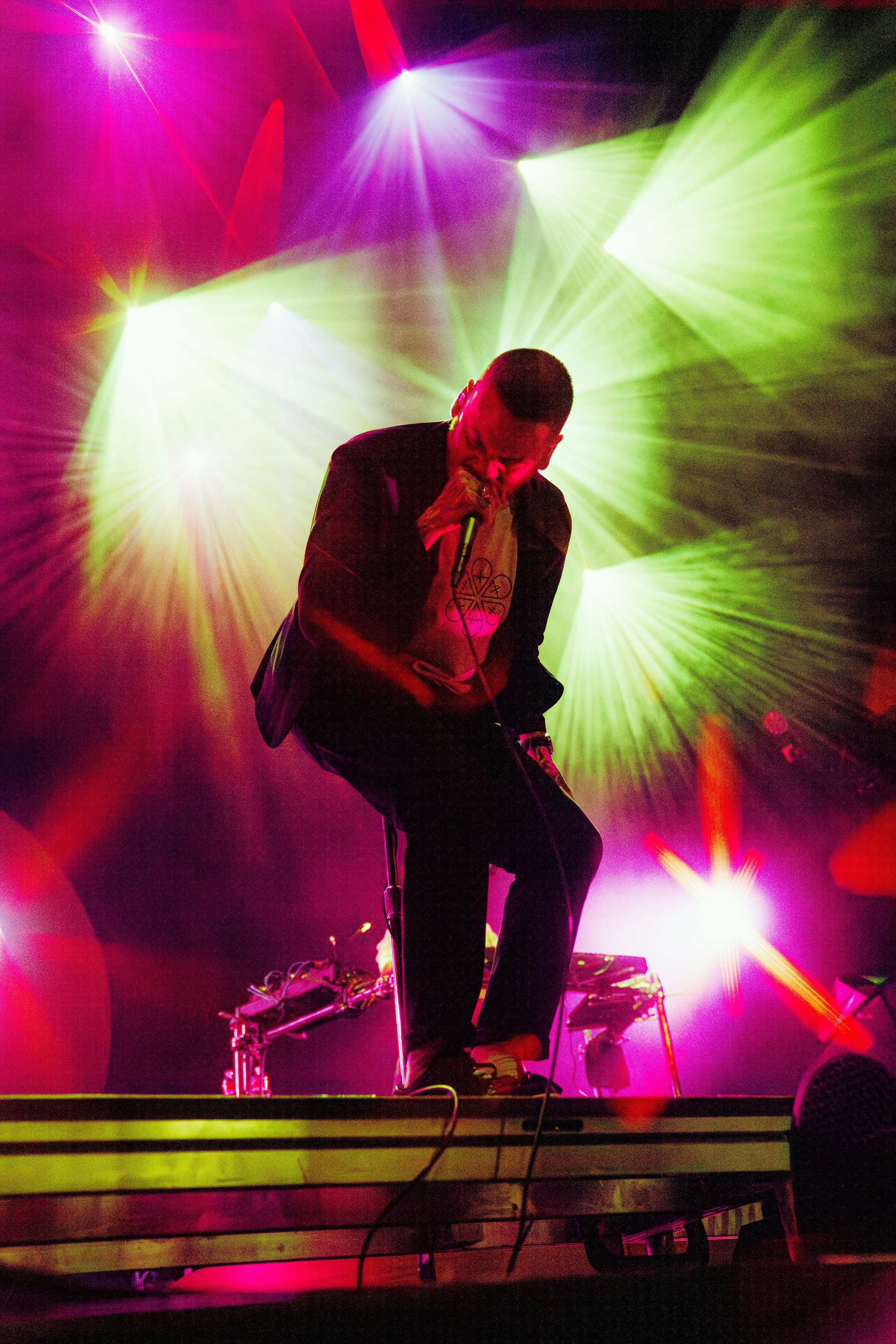 A musician performing on stage with colorful laser lights shining in purple, green, yellow, and red in the background. The performer is bent over, holding a microphone, with a DJ setup and electronic equipment visible on the stage.