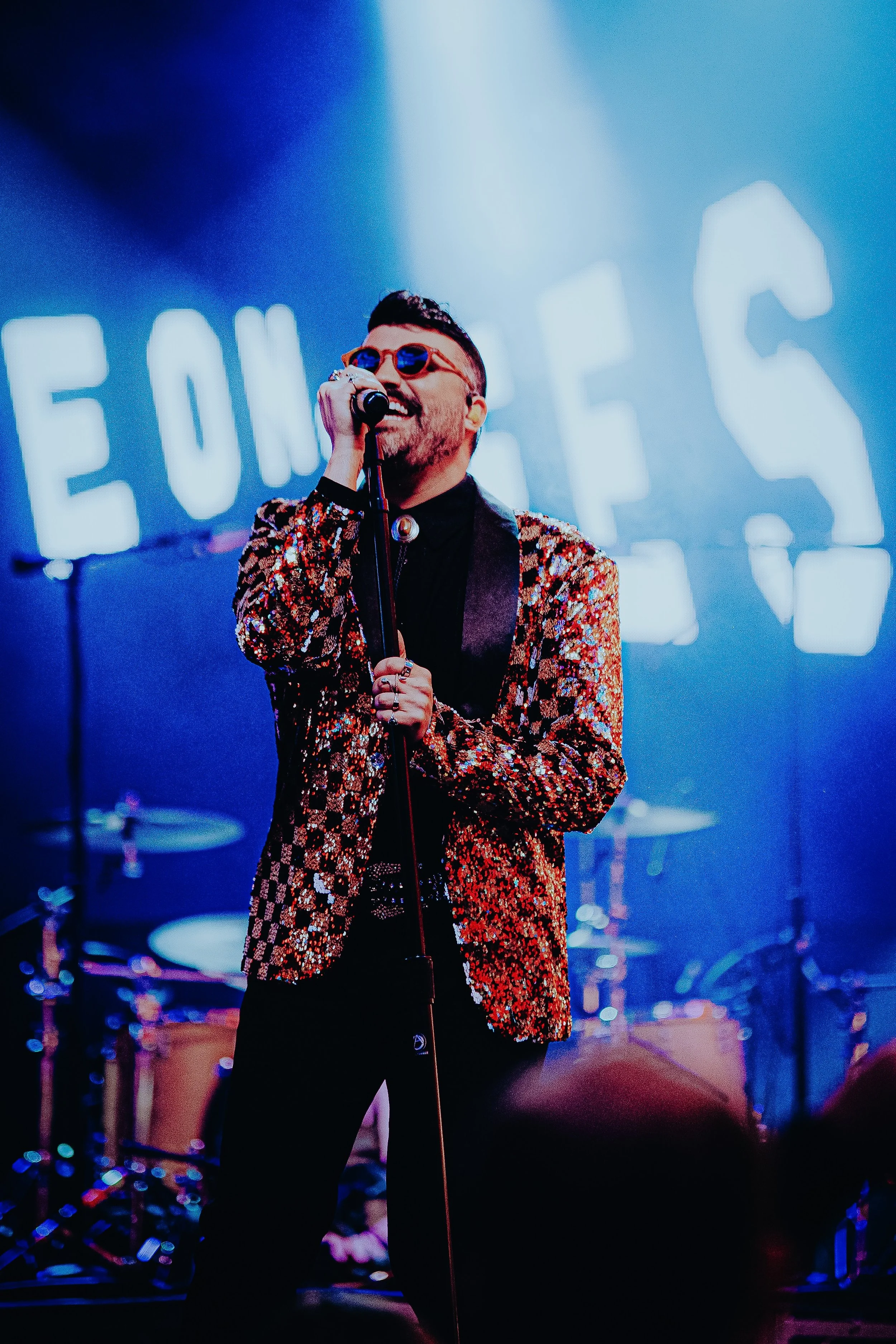A man wearing a sequined blazer and orange sunglasses is singing into a microphone on stage, with a background of musical instruments and a large illuminated sign that reads 'FOUR TAS'.