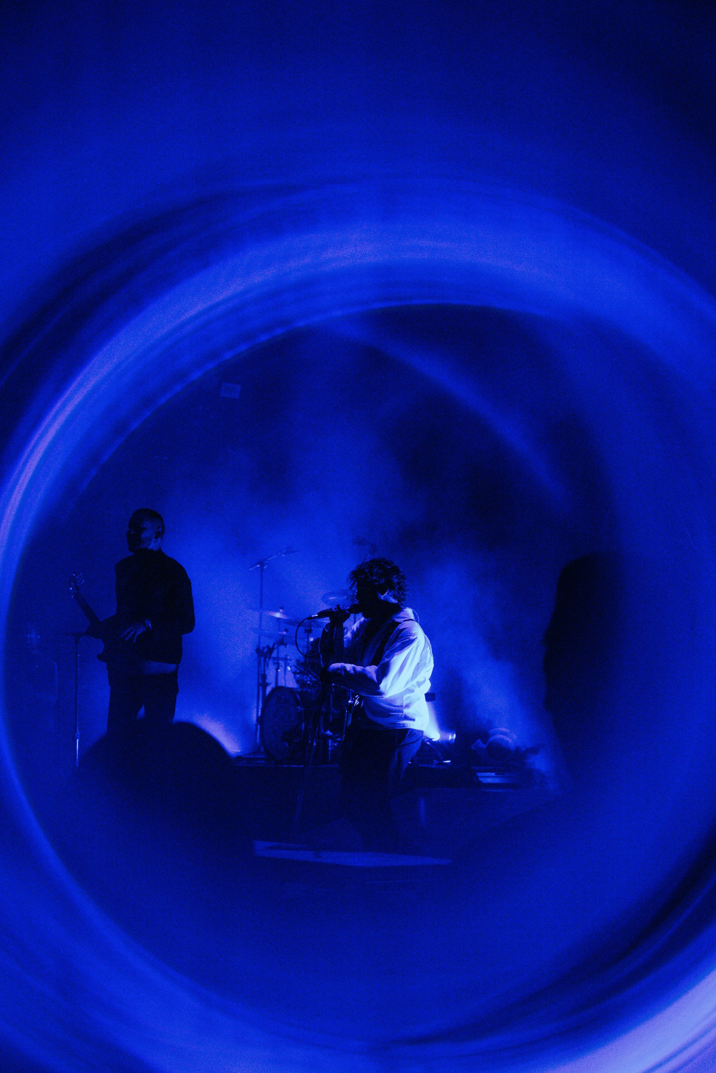 A live musical performance with band members playing instruments on stage, illuminated in blue lighting, with audience silhouettes visible in the foreground.