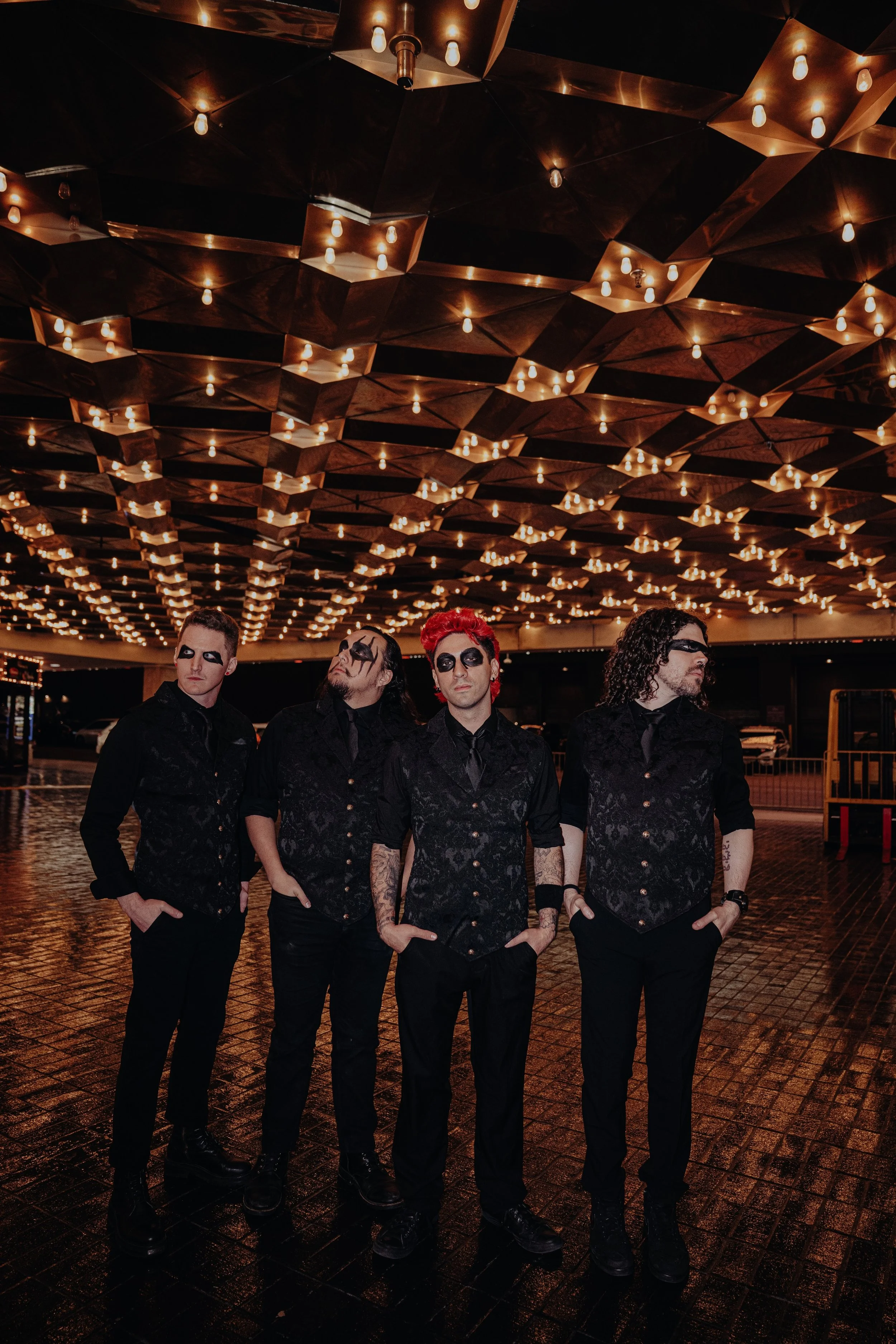 Four men dressed in black with dramatic makeup and tattoos standing under a ceiling with numerous warm orange light bulbs in an indoor space with a wet, reflective floor.