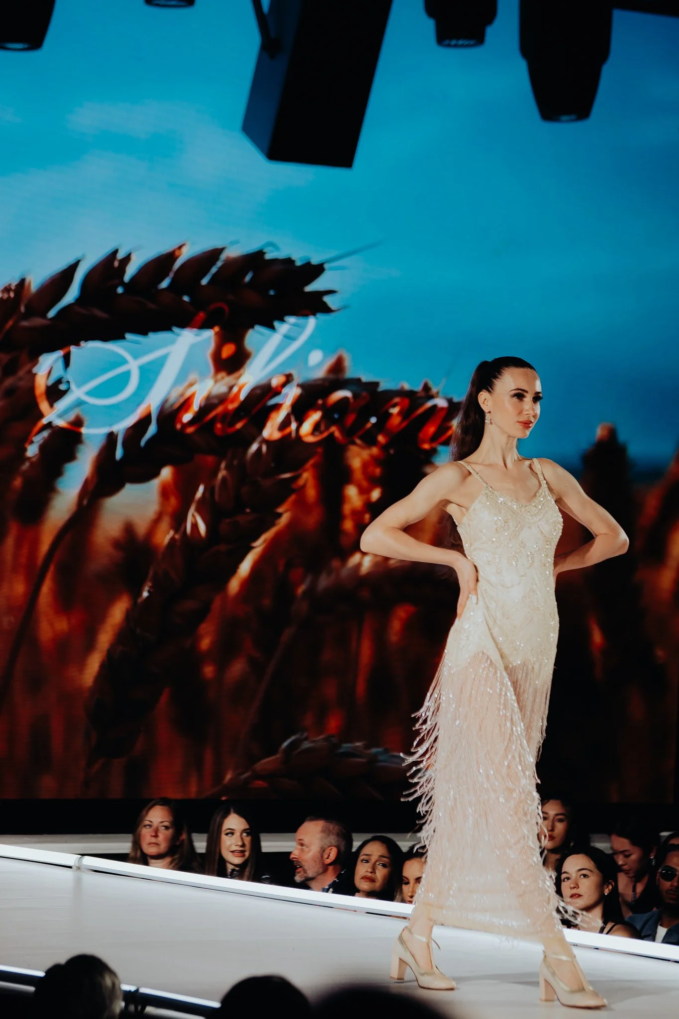 A woman in a beige, embellished dress and high heels walking on a runway during a fashion show, with an audience seated in the background and blue sky visible above.