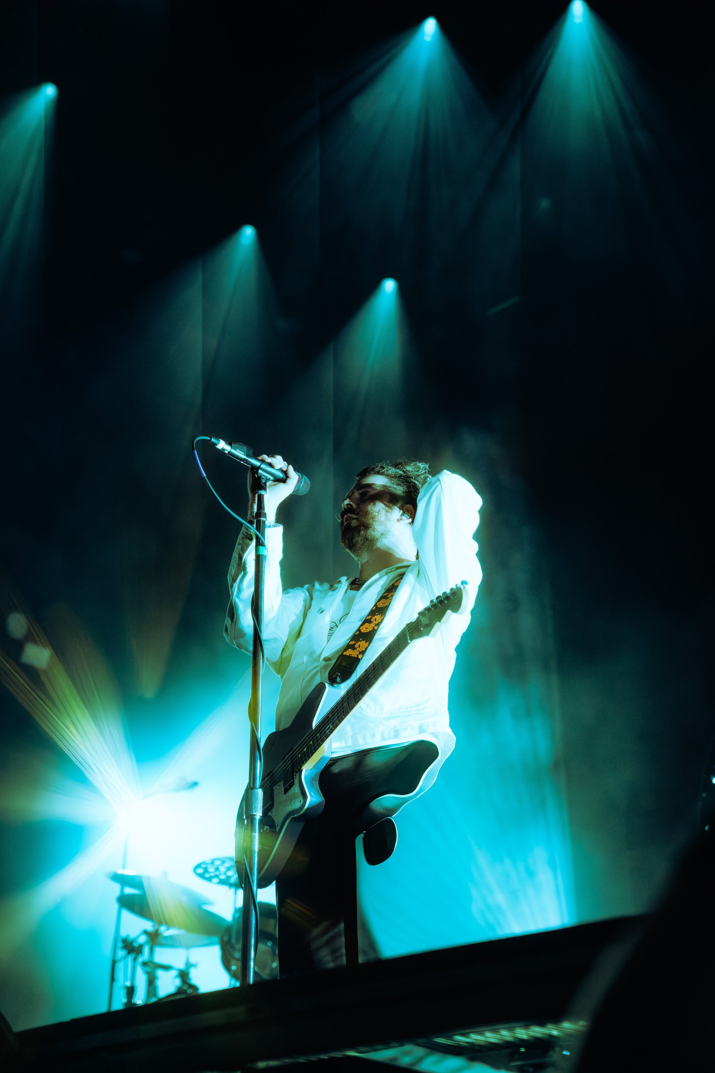 A singer with curly hair wearing white shirt and black pants performing on stage with a guitar, illuminated by blue and green stage lights.