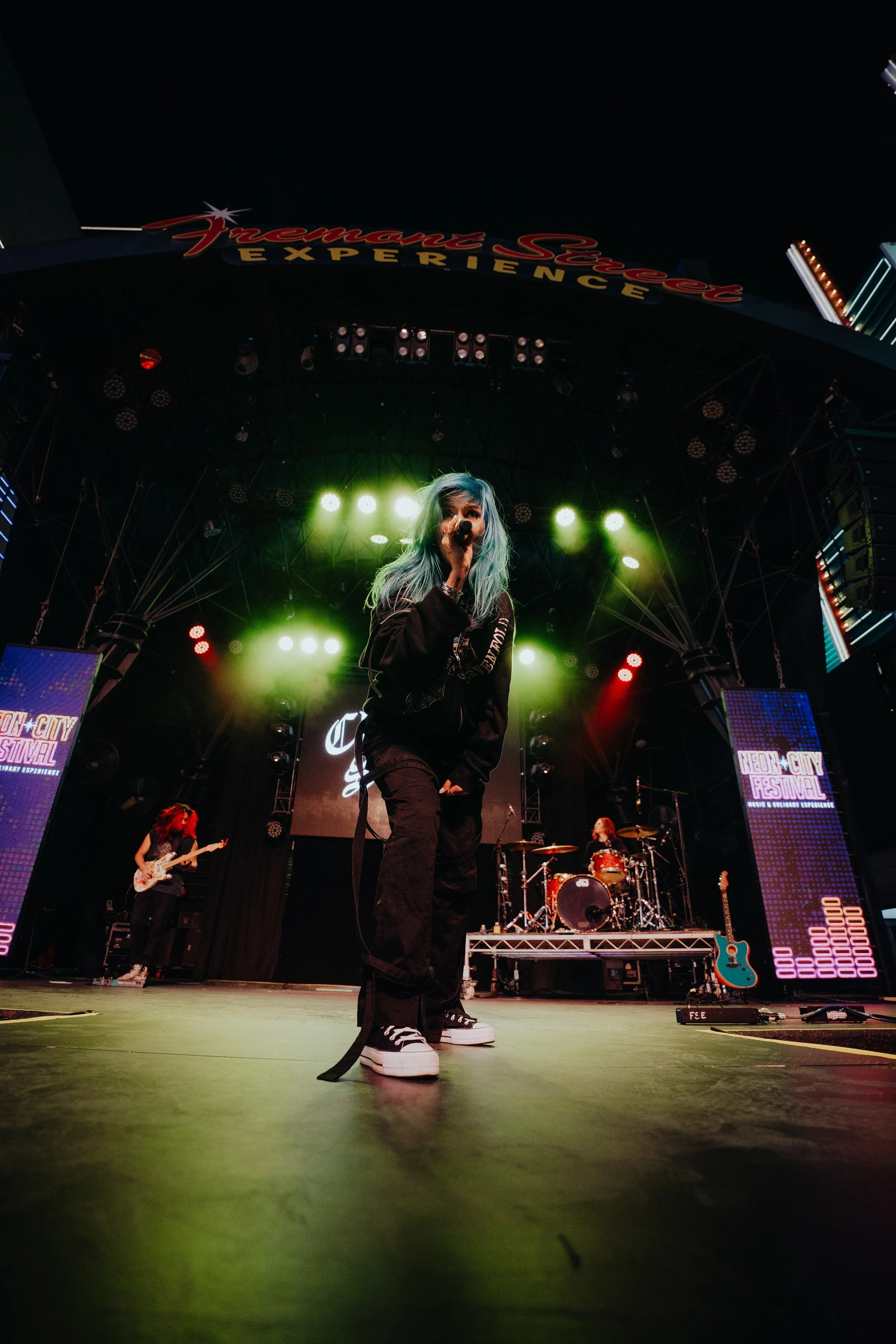 Female lead singer with blue hair performing on stage with green lighting, accompanied by band members playing guitar and drums at a concert at the Neon City Festival.