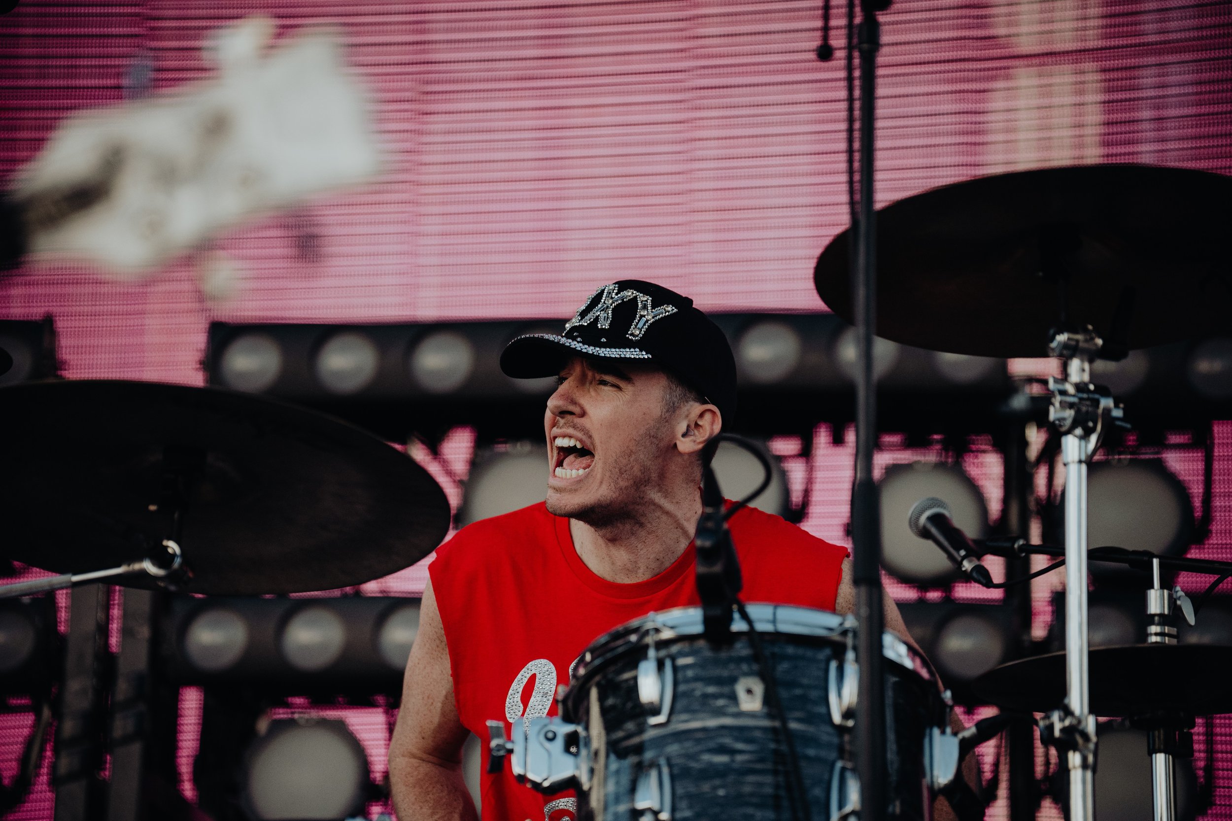 A drummer wearing a black cap with silver embellishments and a red sleeveless shirt playing drums on stage with a pink LED background.