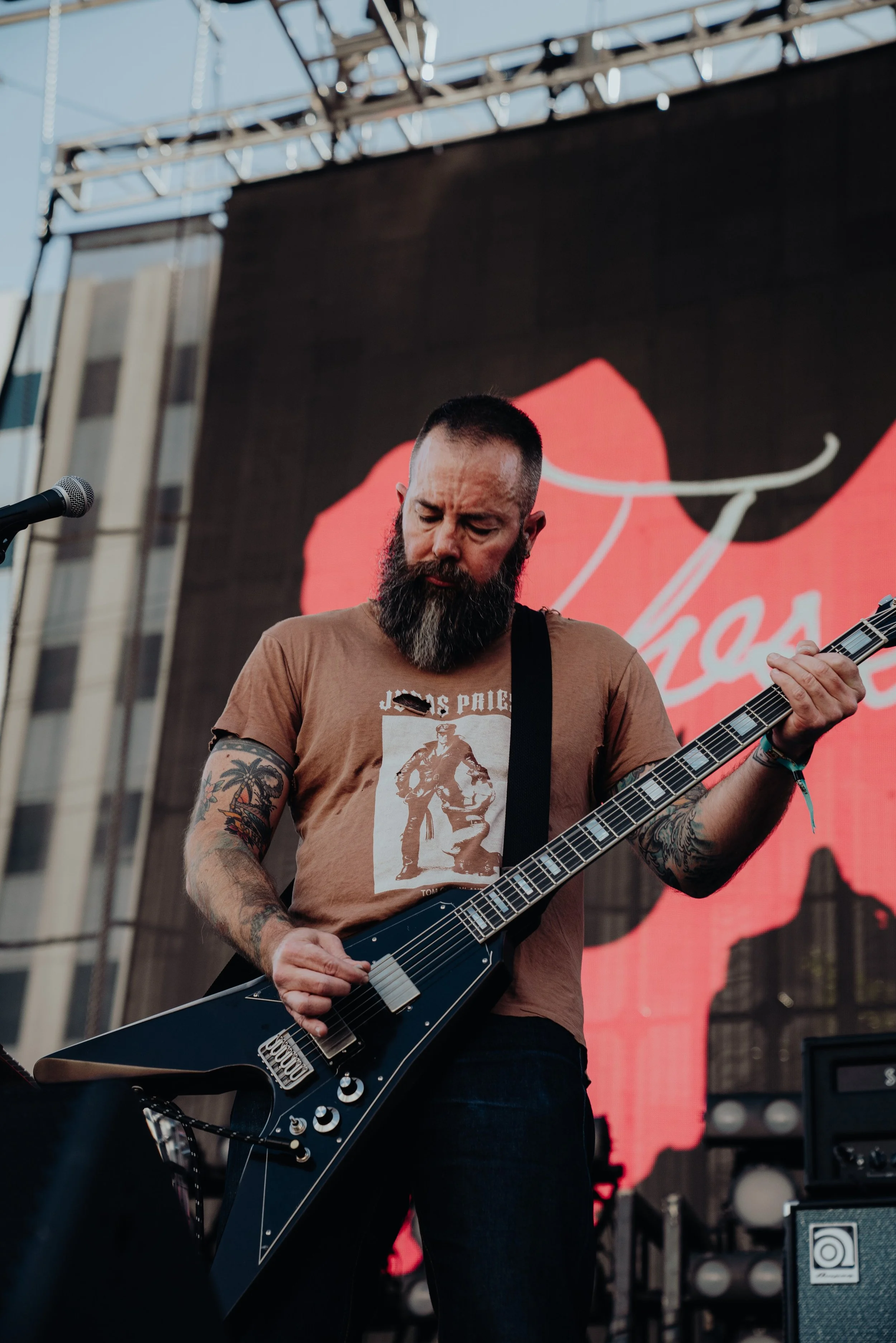 A man with a beard and tattoos playing an electric guitar on an outdoor stage during a live concert. There is a microphone to his left and a digital screen in the background displaying a colorful graphic.
