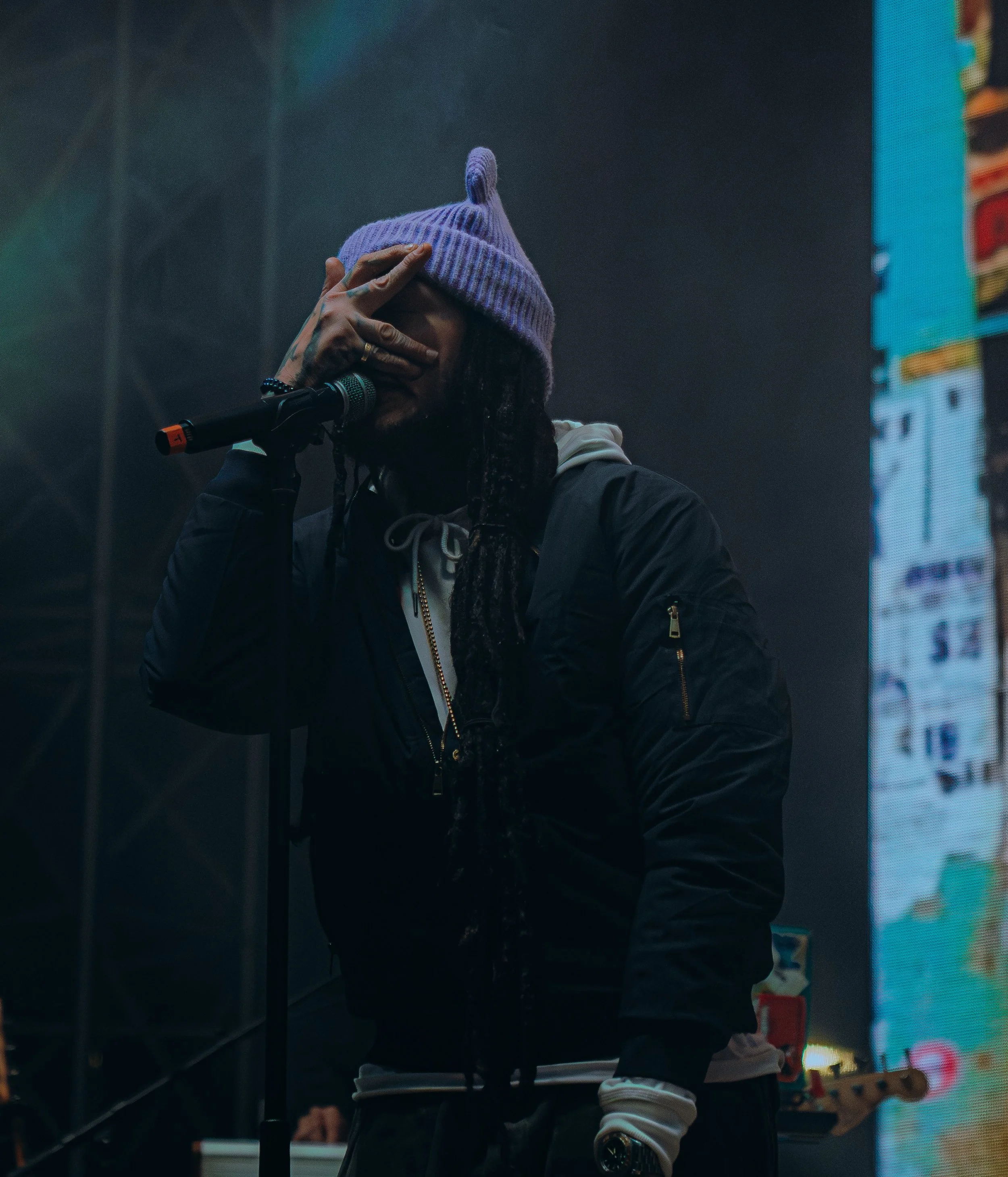 A musician wearing a purple knit beanie is performing on stage, covering their face with their hand. They are dressed in dark clothing with a watch on their wrist, standing in front of a microphone, in front of a colorful LED screen.
