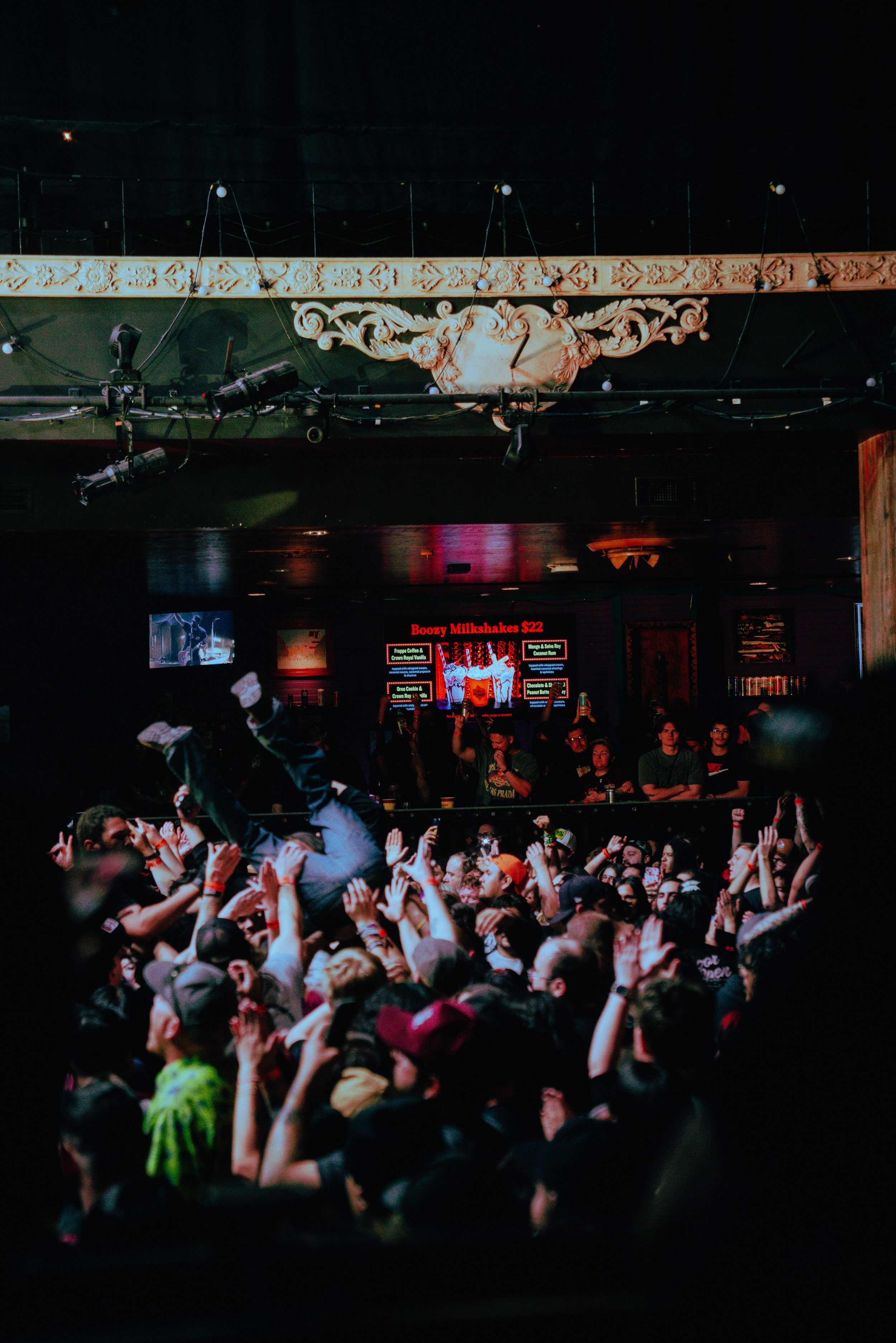 A crowded concert or party scene with people dancing, raising their hands, and crowd surfing. There is a stage with lighting and a screen advertising drinks, with a sign saying 'Boozy Milkshakes $22'.