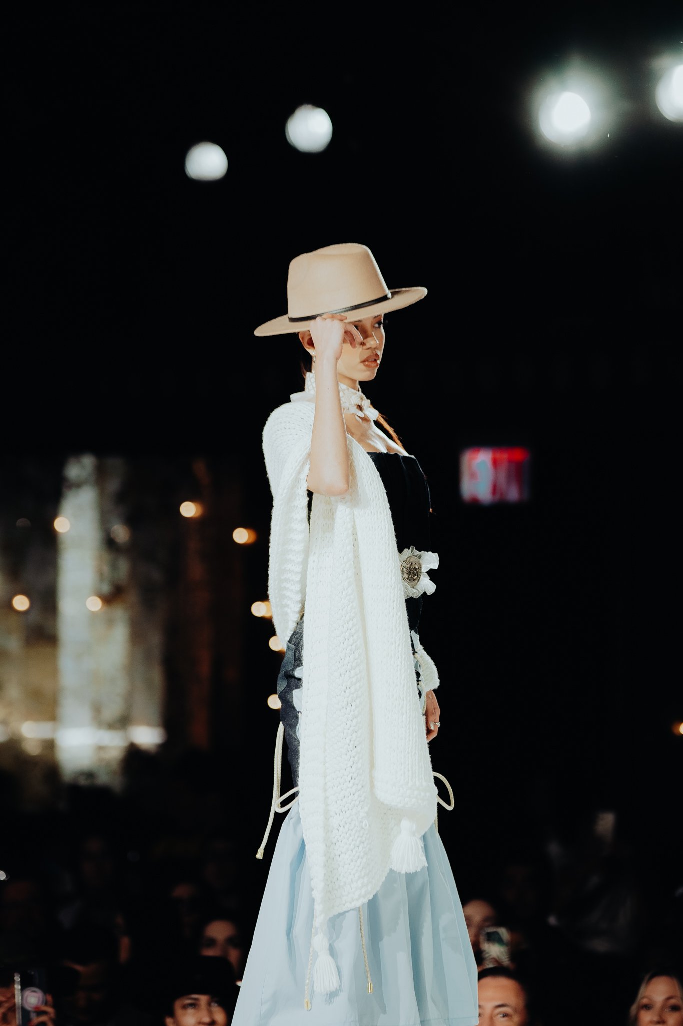 A woman on a runway wearing a large beige hat, a white textured outerwear, and a flowing light blue skirt, with an audience visible in the background.