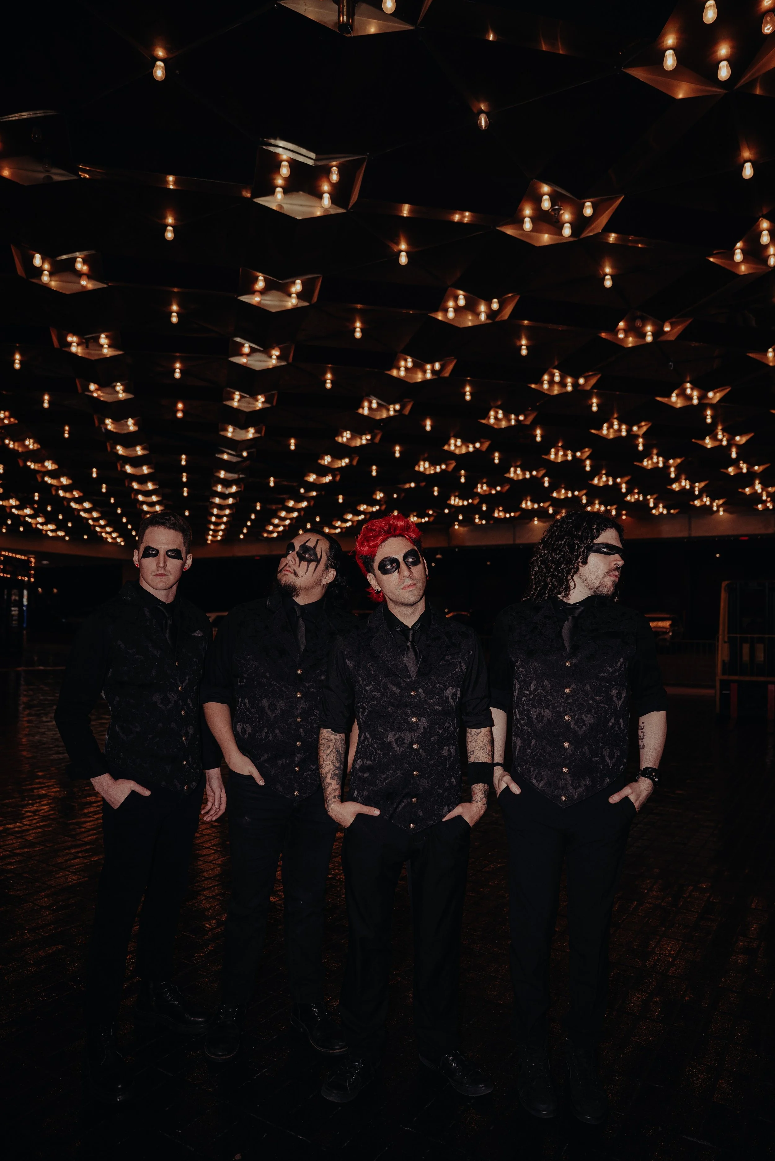 Four men dressed in black with dark makeup pose under a ceiling with numerous small pendant lights in a dark setting.