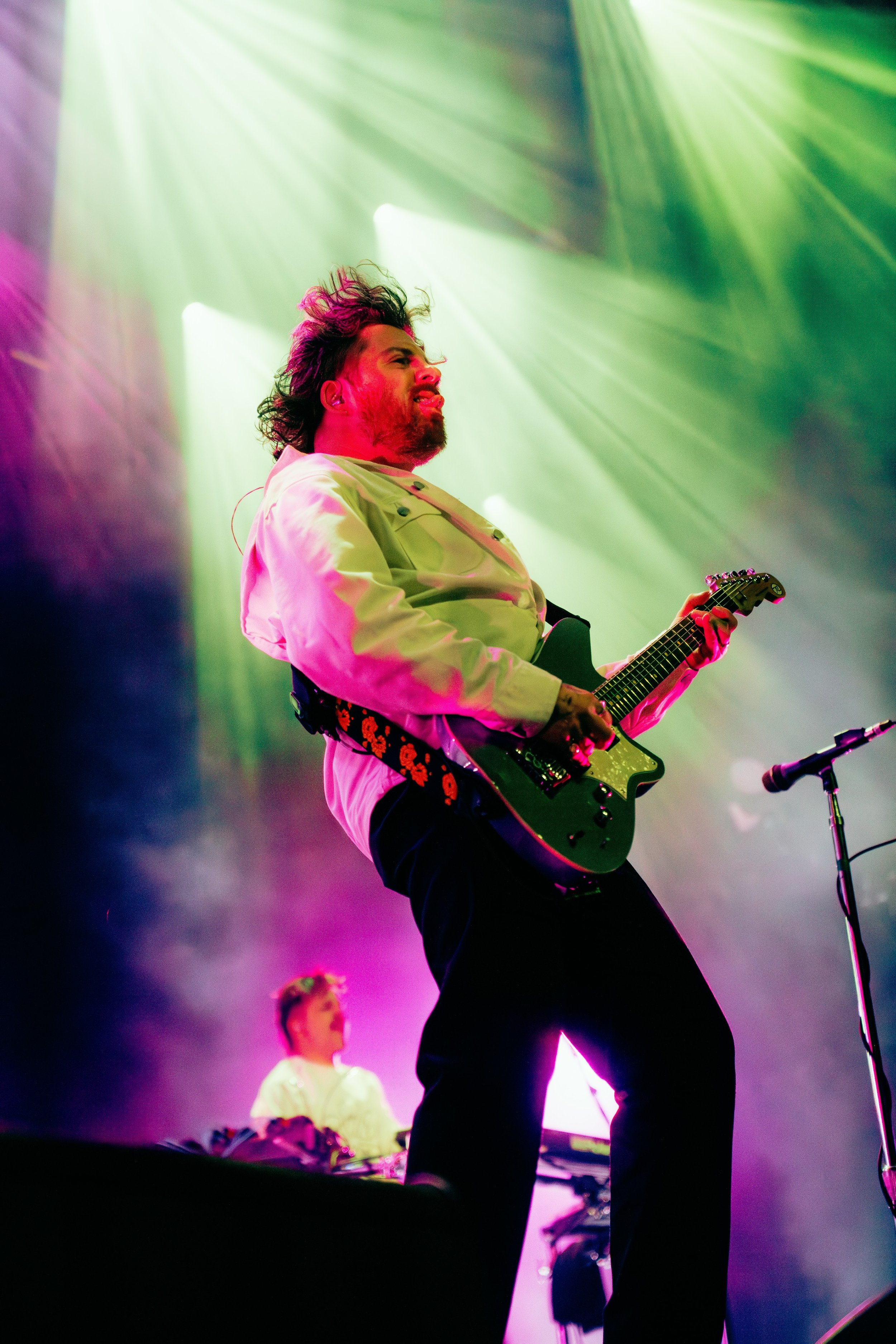 A male guitarist with dark hair and beard performing on stage with colorful lighting, wearing a light green jacket and black pants, with another musician visible in the background.