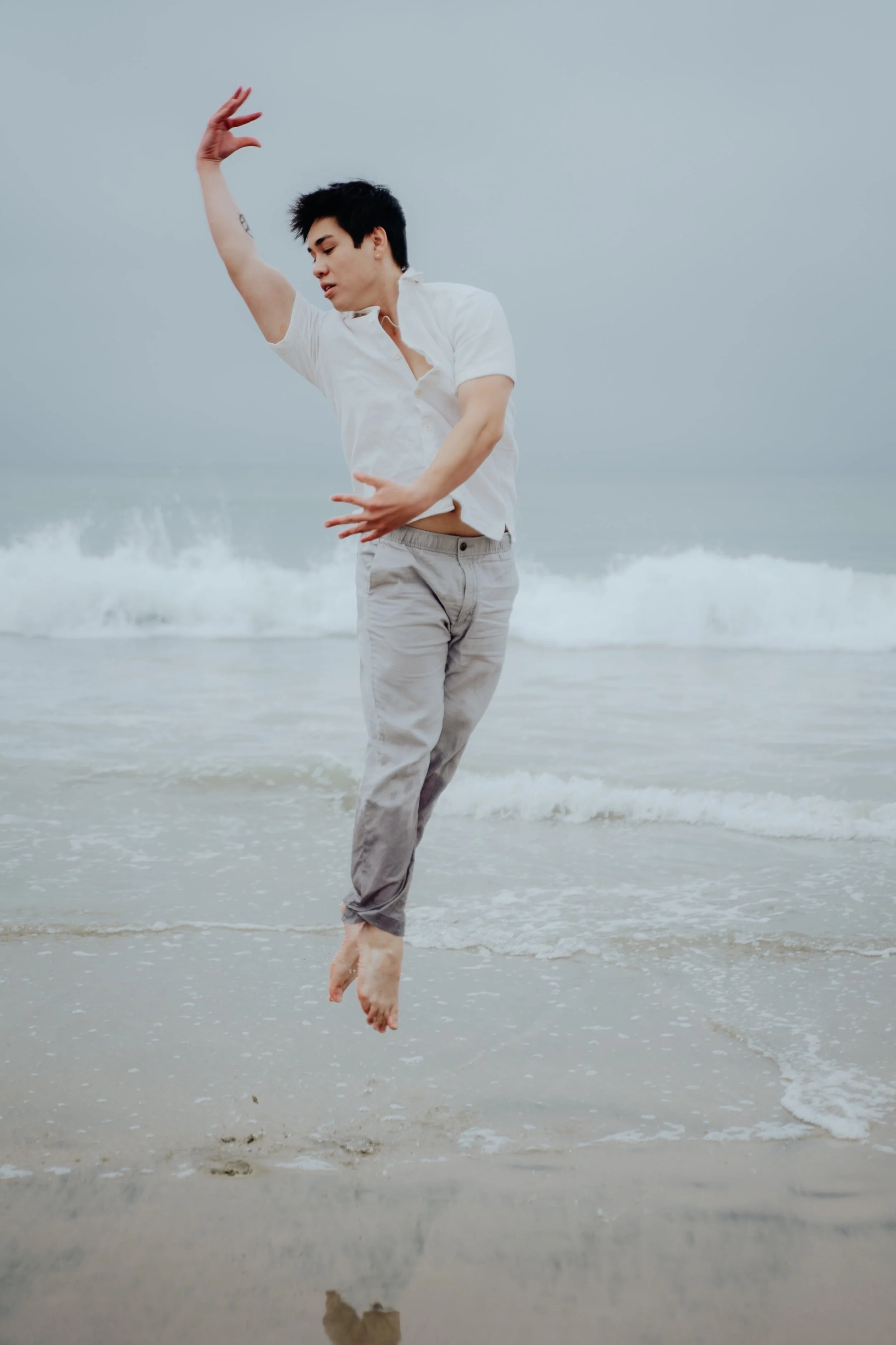 Person dancing on the beach with their feet in the water, wearing a white shirt and beige pants, with waves crashing behind them.