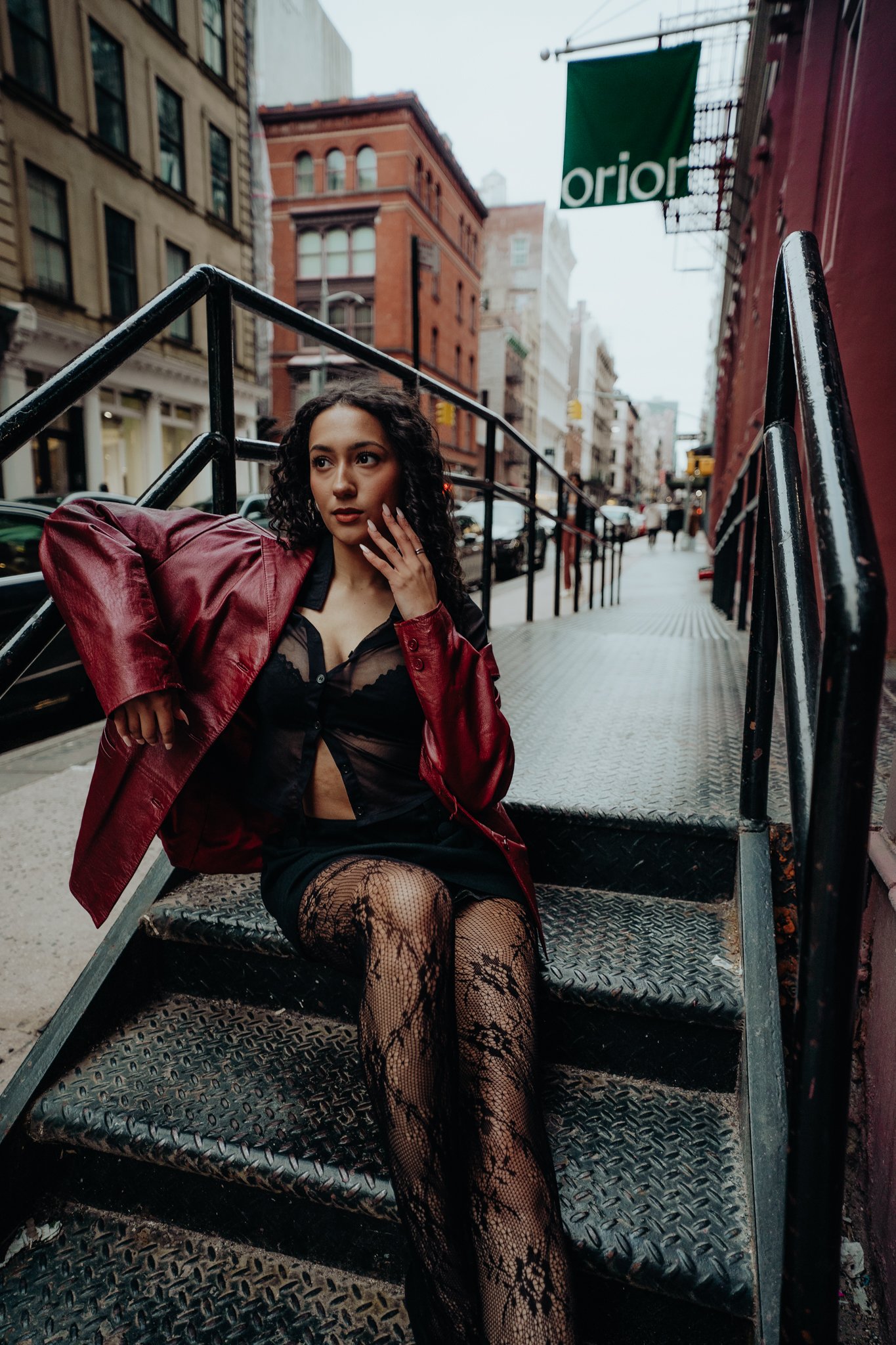 A woman with curly hair sitting on metal stairs in an urban street, wearing a red jacket, black lace dress, and fishnet stockings, with buildings and cars in the background.