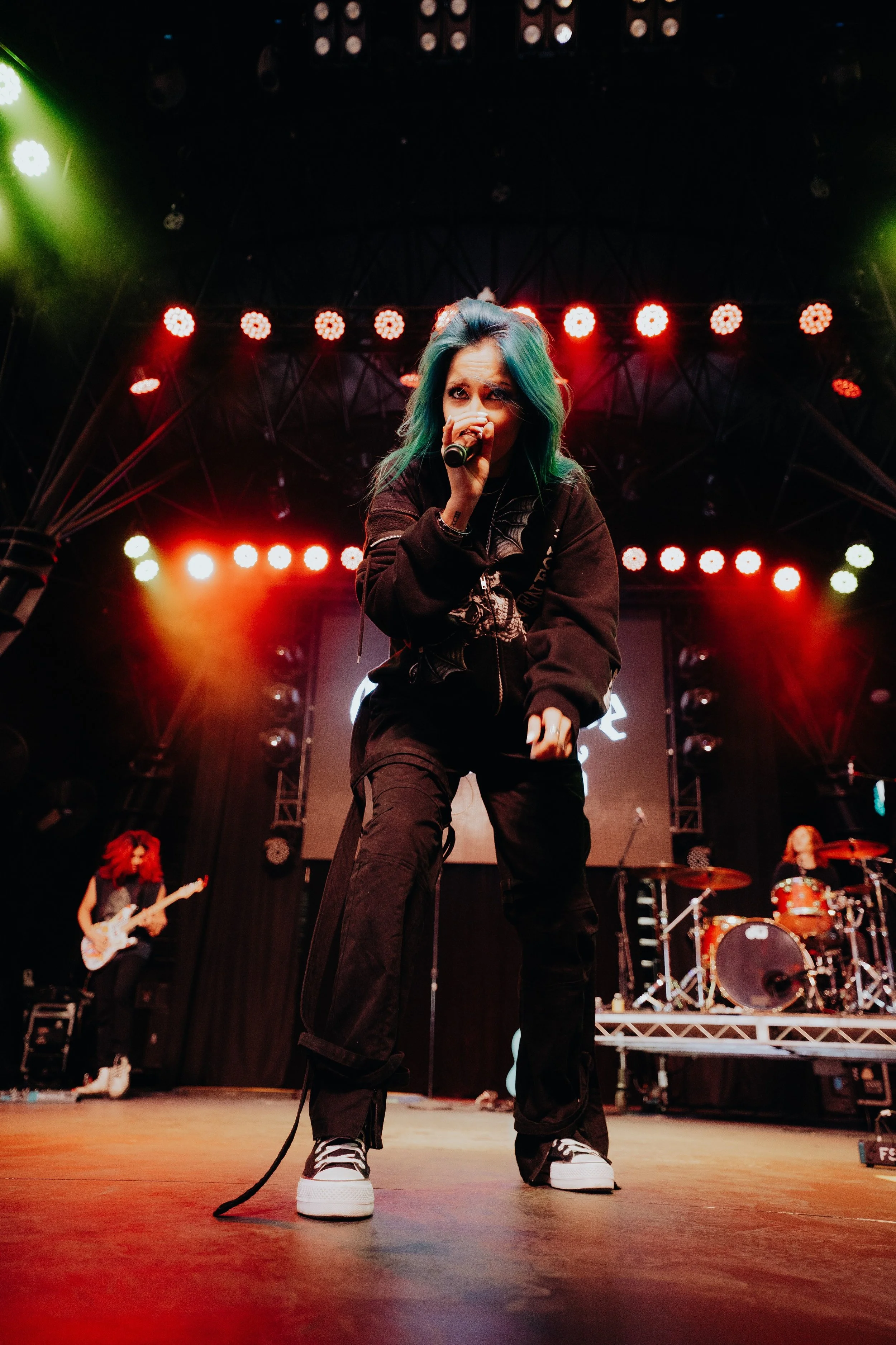 Female singer with green and blue hair performing on stage with band members playing guitar and drums, stage lights and screens in background.