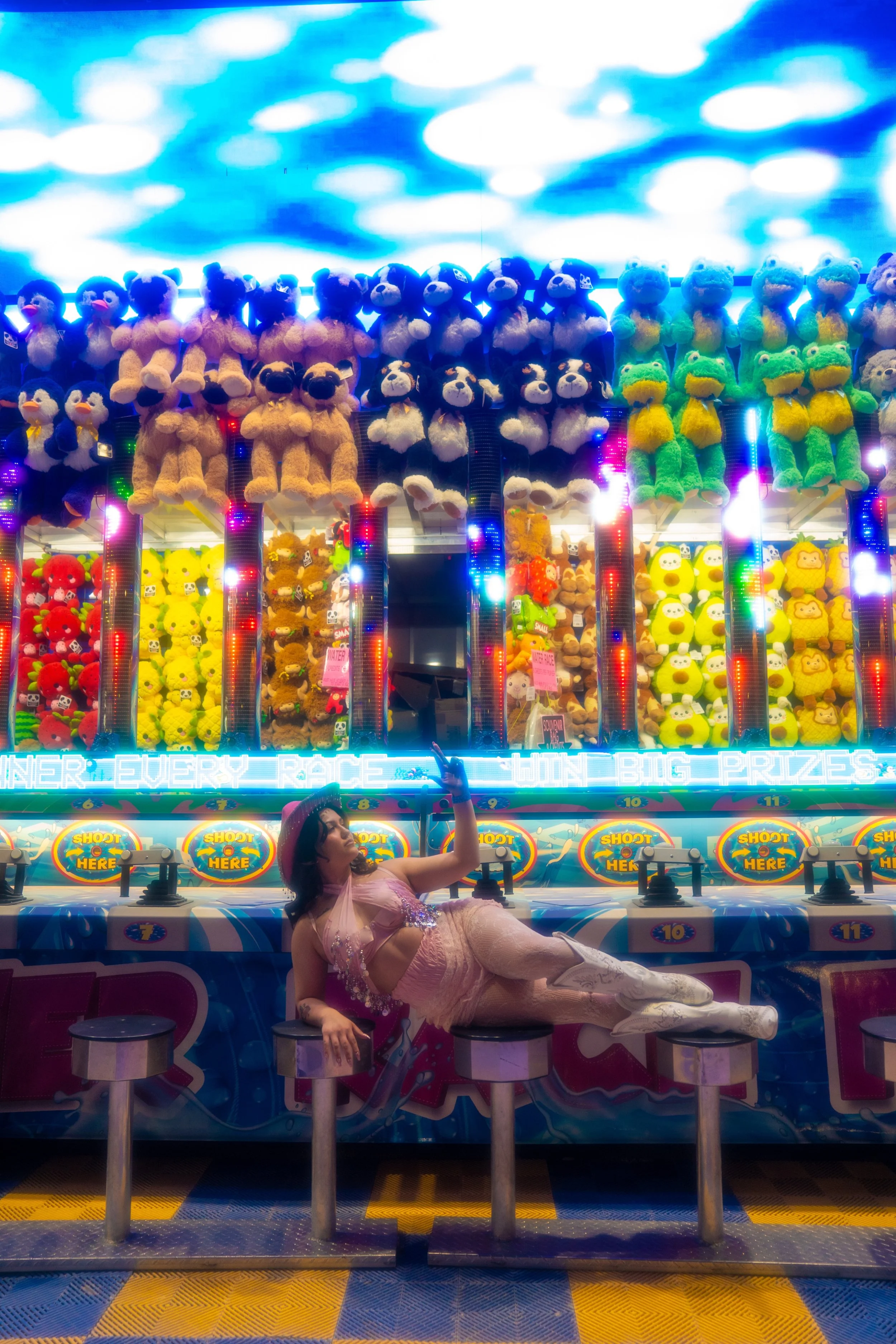 Fairground game booth with plush toys including teddy bears, frogs, and ducks hanging above. A woman in a lace dress reclining on a carnival game counter, holding a balloon, with colorful lights and a patterned floor.