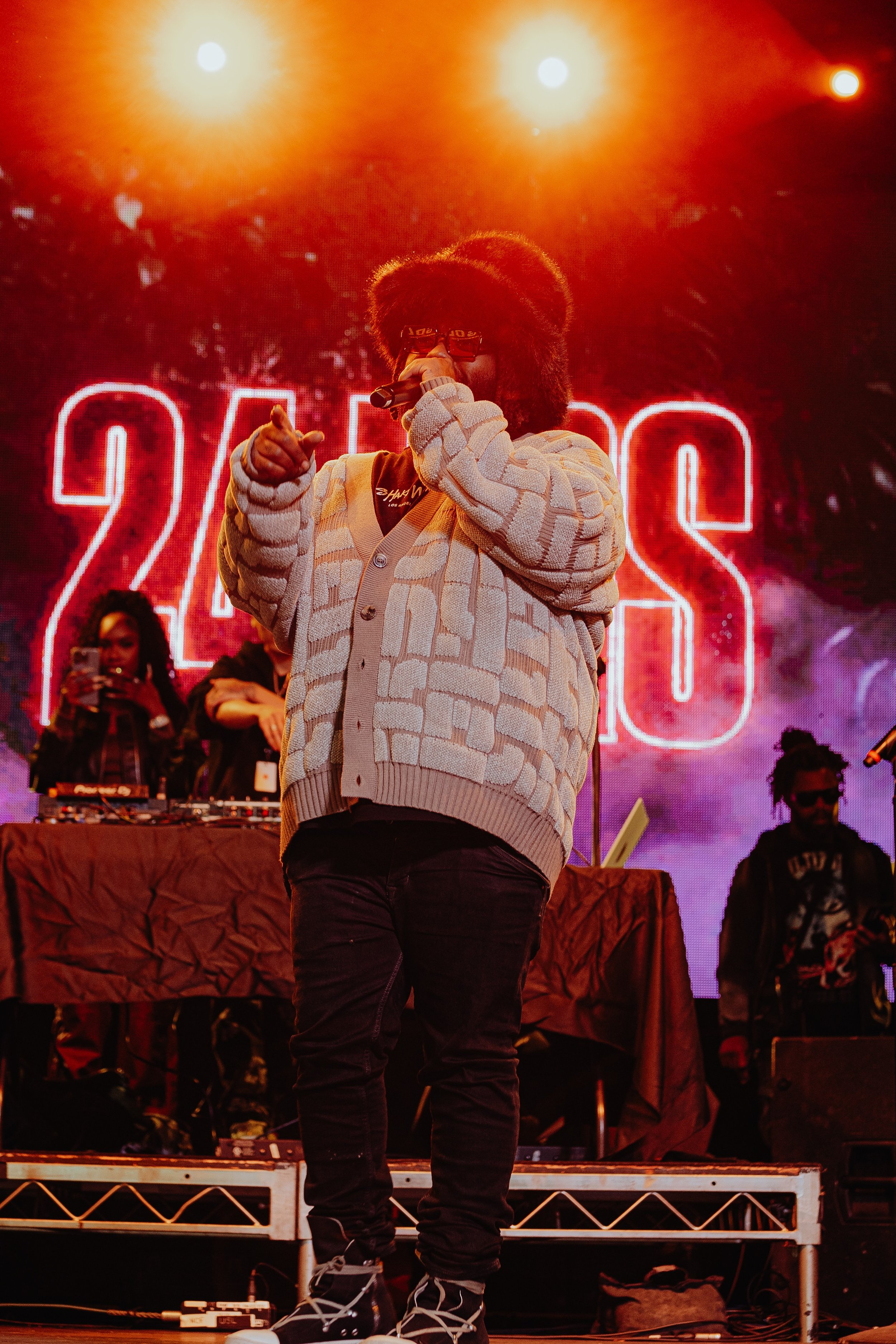 A performer in glasses and a patterned beige jacket singing into a microphone on stage, with a large red '2023' sign in backlit in red lights and an audience member taking photos.