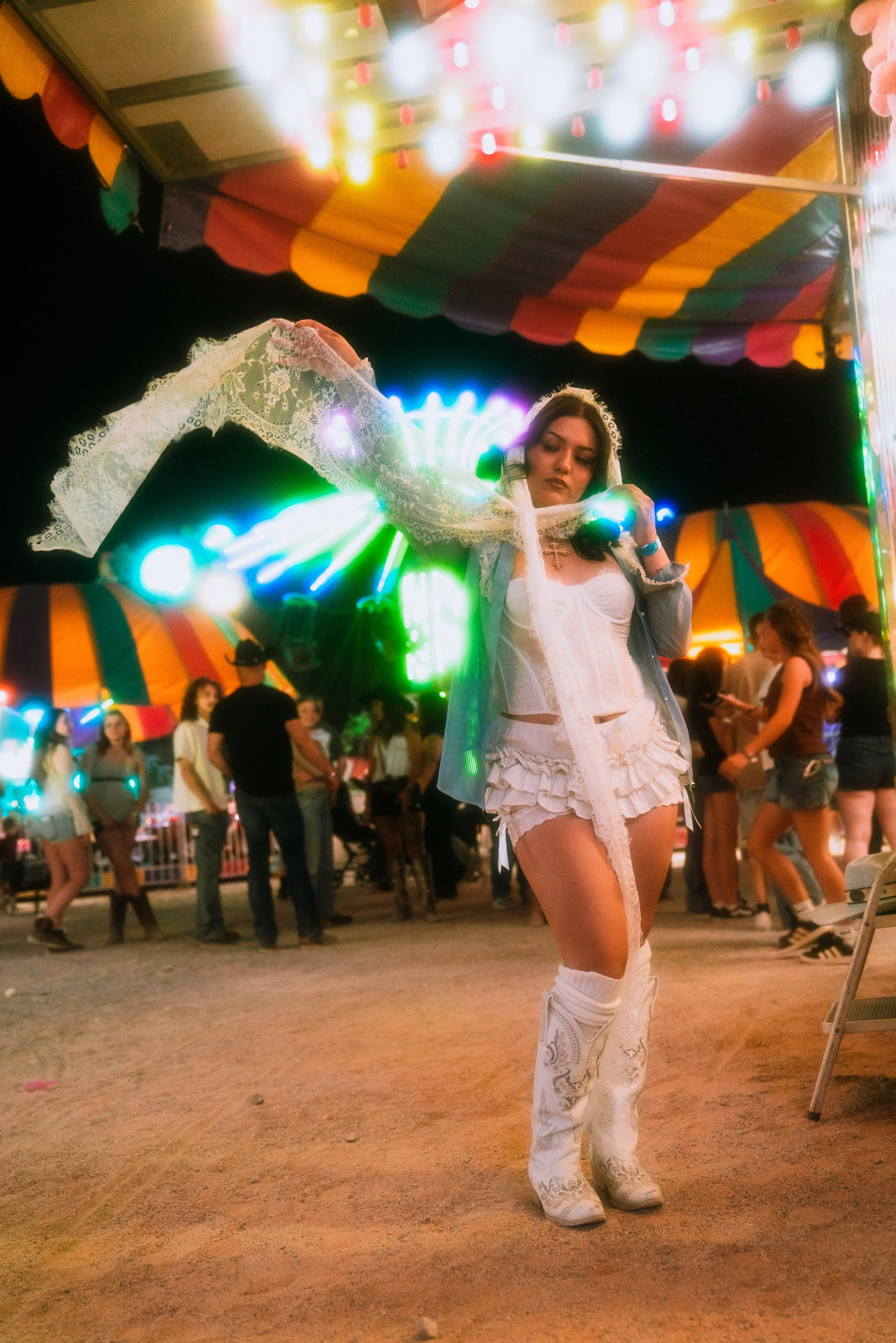 A woman dressed in a white cowboy outfit, including a lace shirt, ruffled shorts, and cowboy boots, is dancing at a fair at night under colorful lights and decorations. There are other people in the background enjoying the fair.