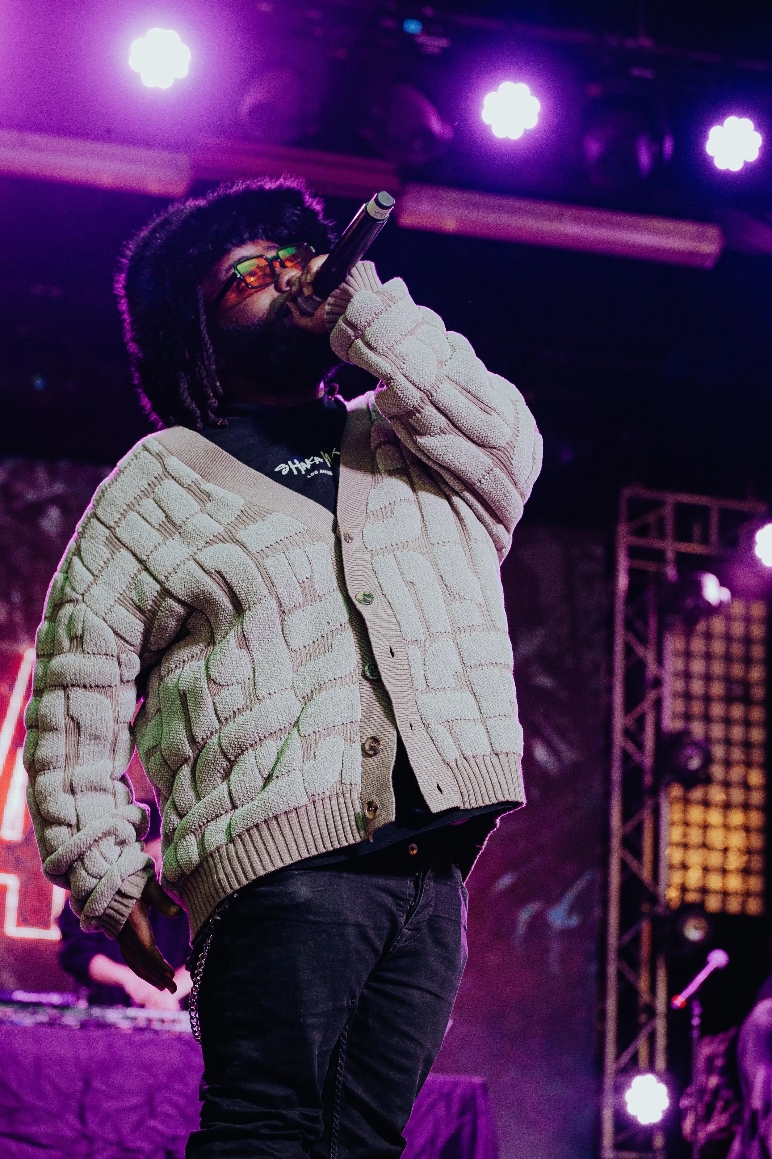 A person singing into a microphone on stage, wearing glasses, a patterned cardigan, and dark pants, with stage lights in the background.