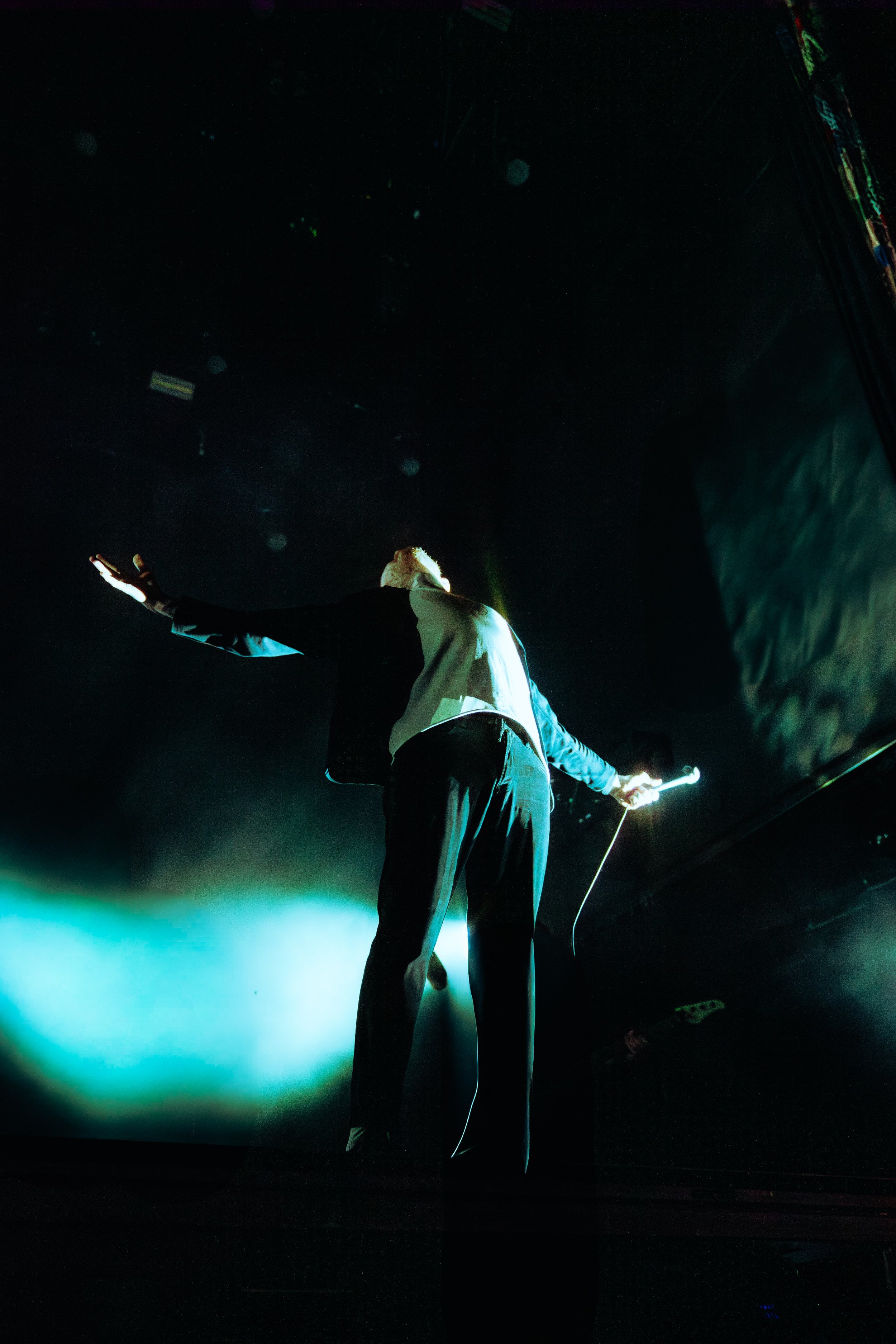 Person in dark clothing on stage, reaching out with hands, illuminated by a greenish light, in a dark, atmospheric setting.