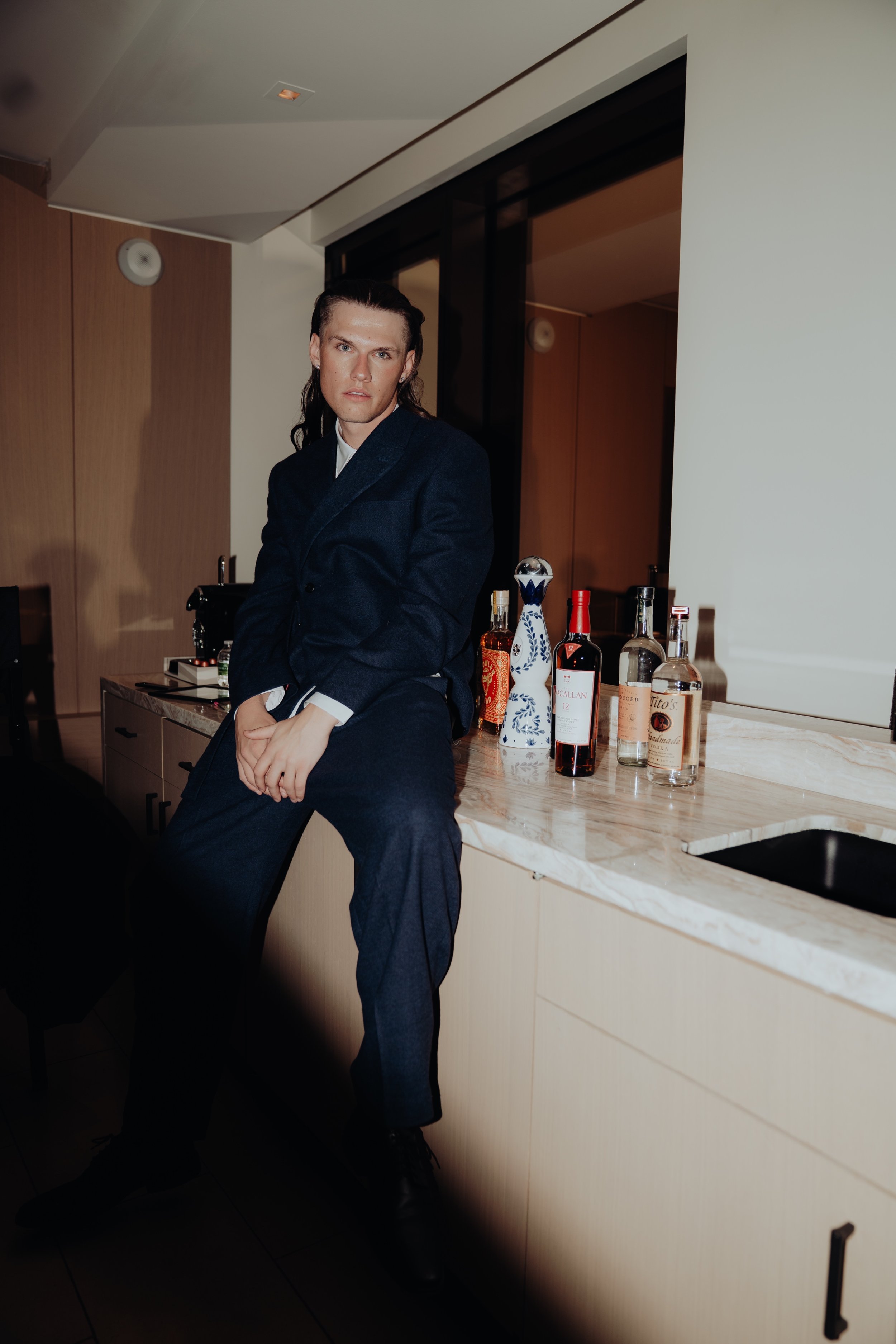 A man with long dark hair, dressed in a dark suit, sitting on a kitchen counter with a marble top. Behind him are various bottles of alcohol and a decorative vase, in a modern home setting.