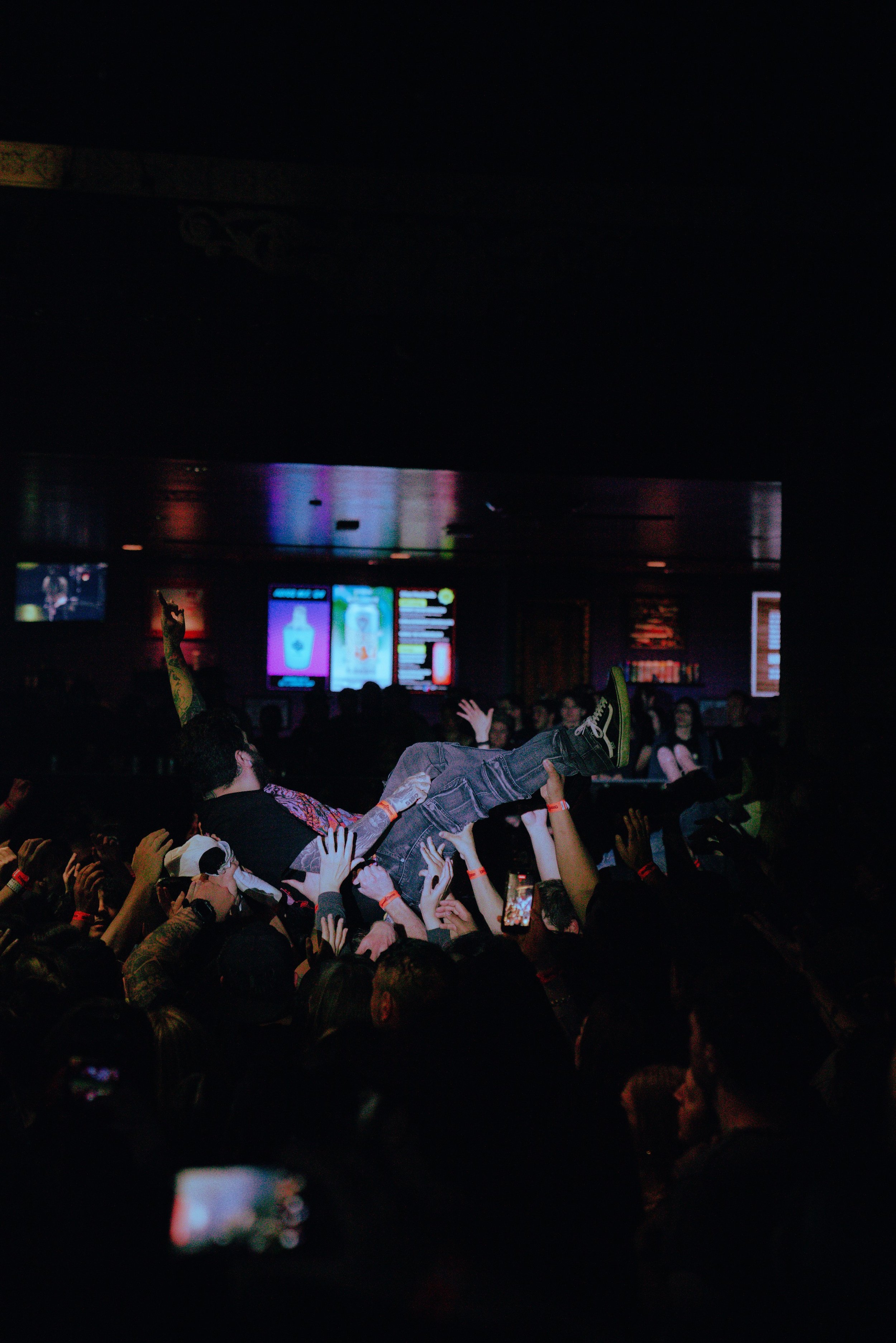 Crowd at concert with person crowd surfing, audience raising hands, and digital displays in the background.