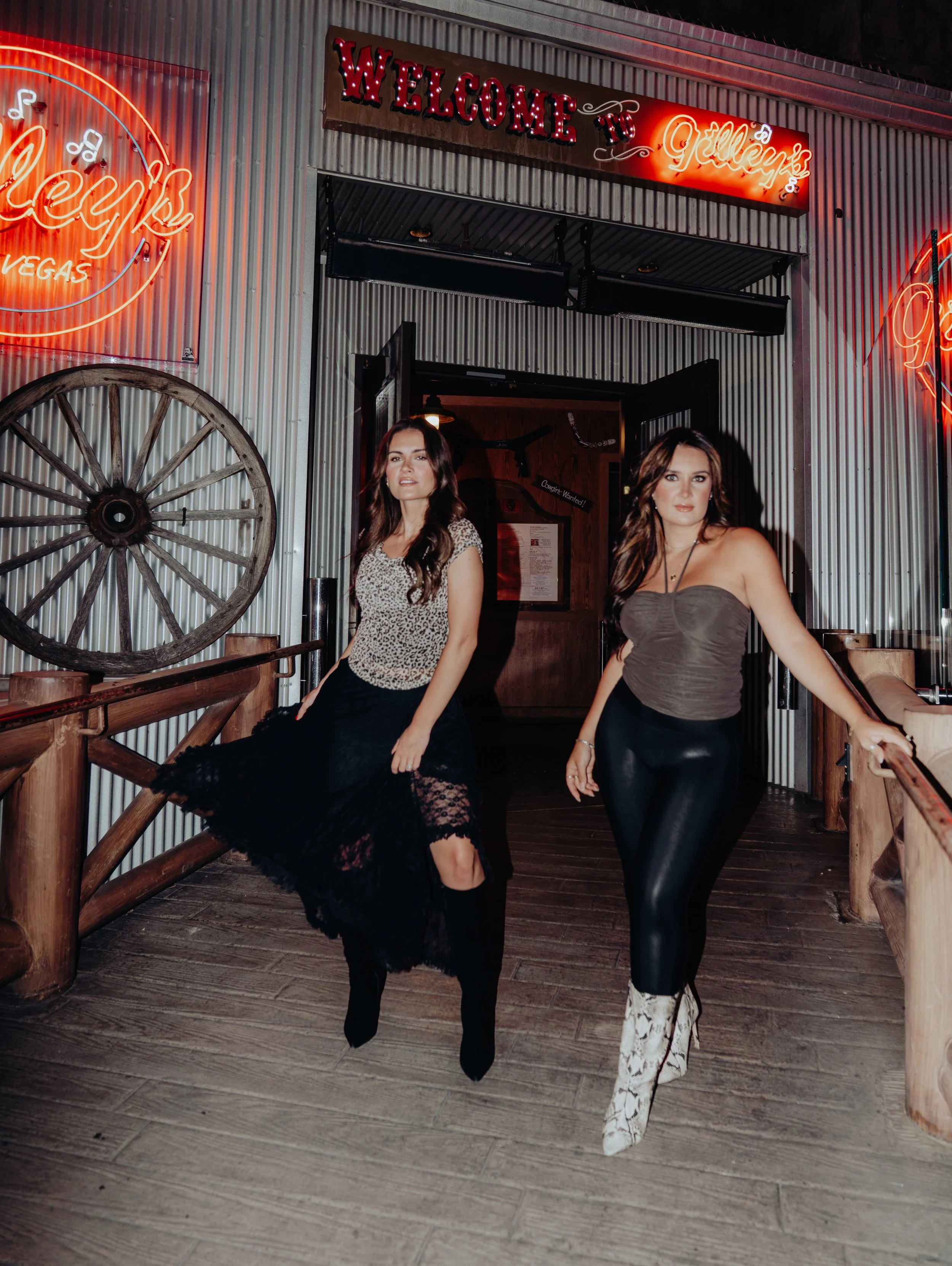 Two women standing in front of the entrance to a restaurant or bar, with neon signs and decorative elements in a western or rustic style, including a wagon wheel and wooden railing.