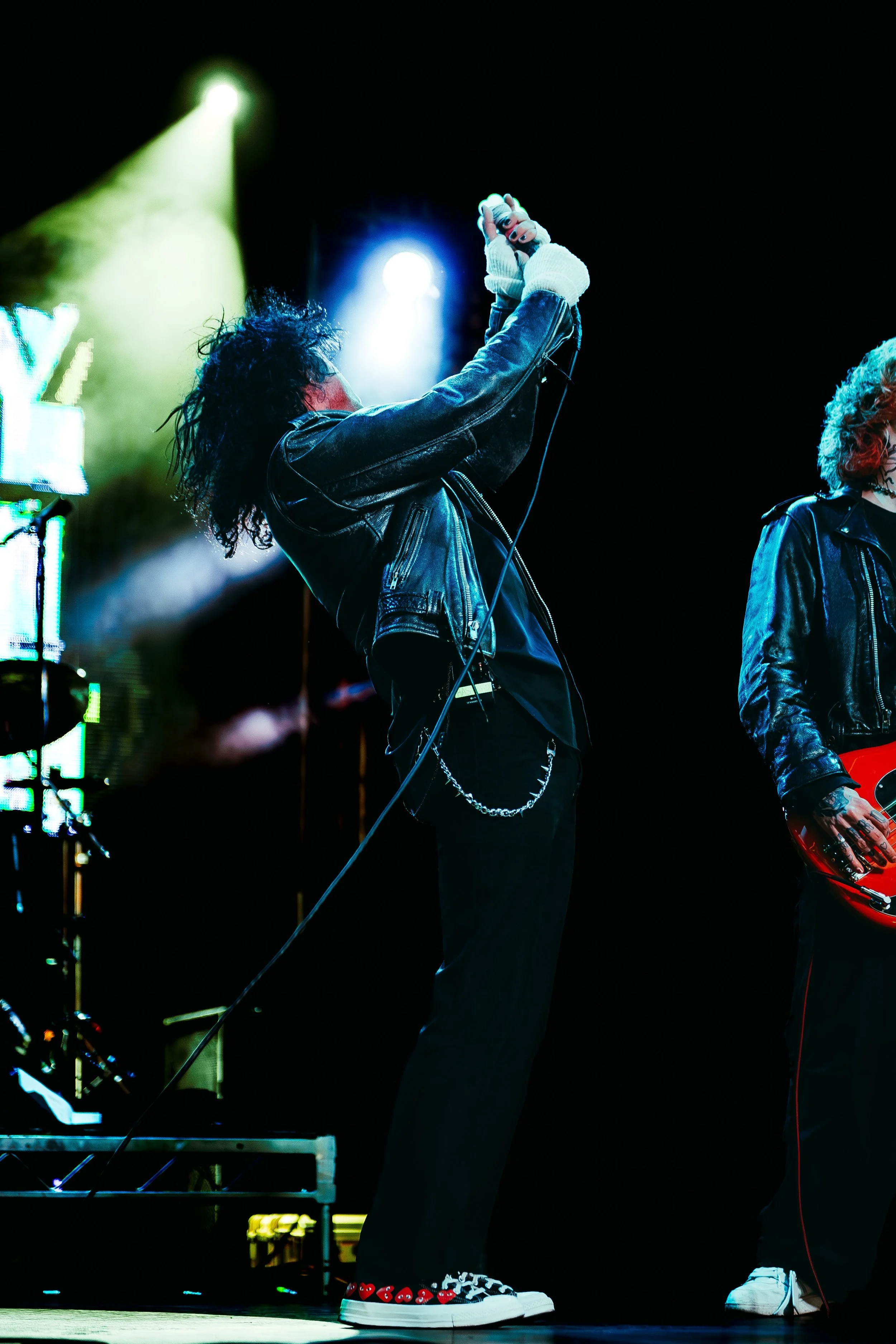 A performer on stage wearing a black leather jacket, black pants, and sneakers with heart designs, holding a microphone, with audio equipment and stage lights in the background.